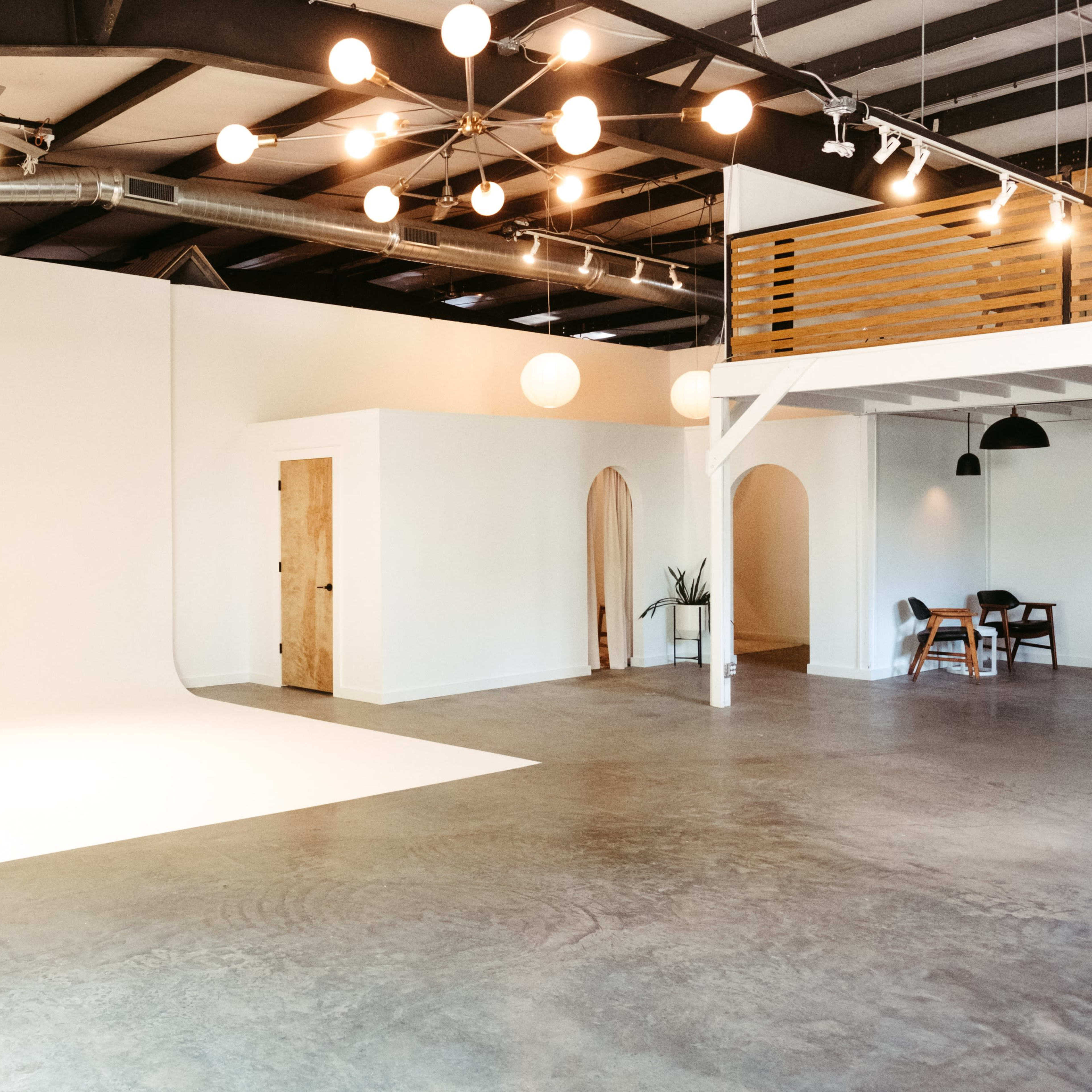 A spacious, well-lit room with a seamless white backdrop, two doors, a seating area, and a mezzanine level with wooden railings.