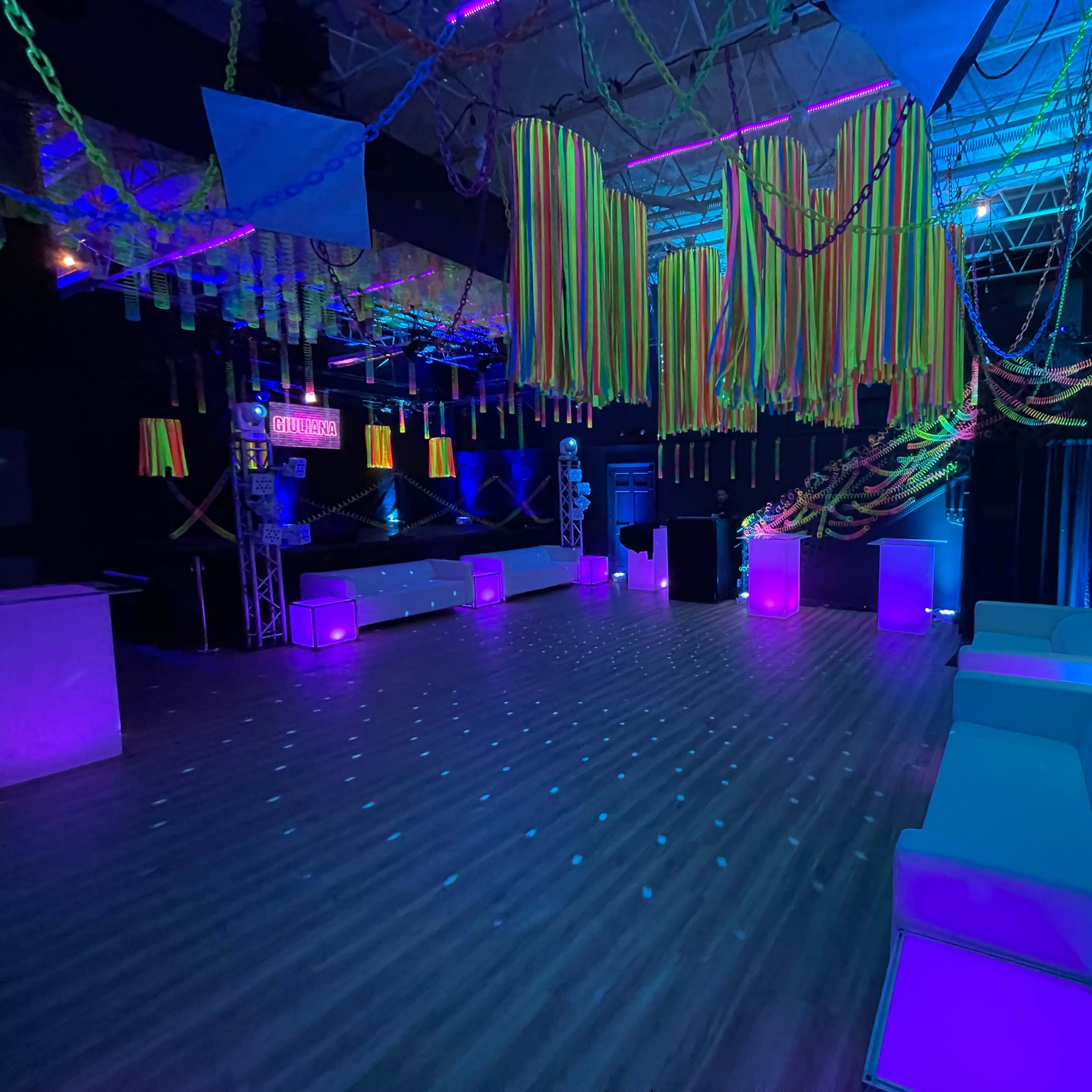 A dimly lit event space decorated with colorful streamers and neon accents, featuring white sofas and illuminated tables.