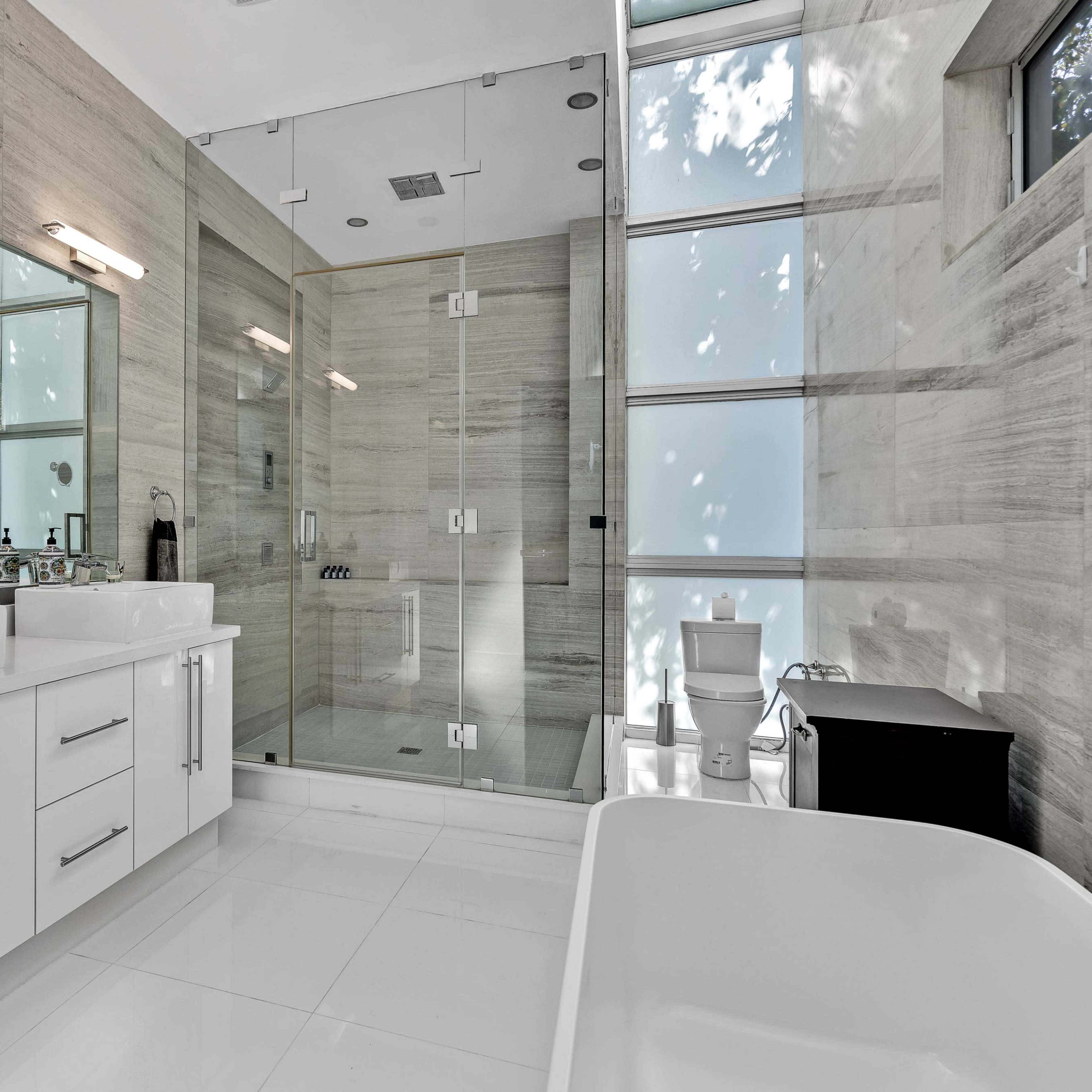 A modern bathroom featuring dual sinks, a glass-enclosed shower, a freestanding bathtub, and large windows allowing natural light.