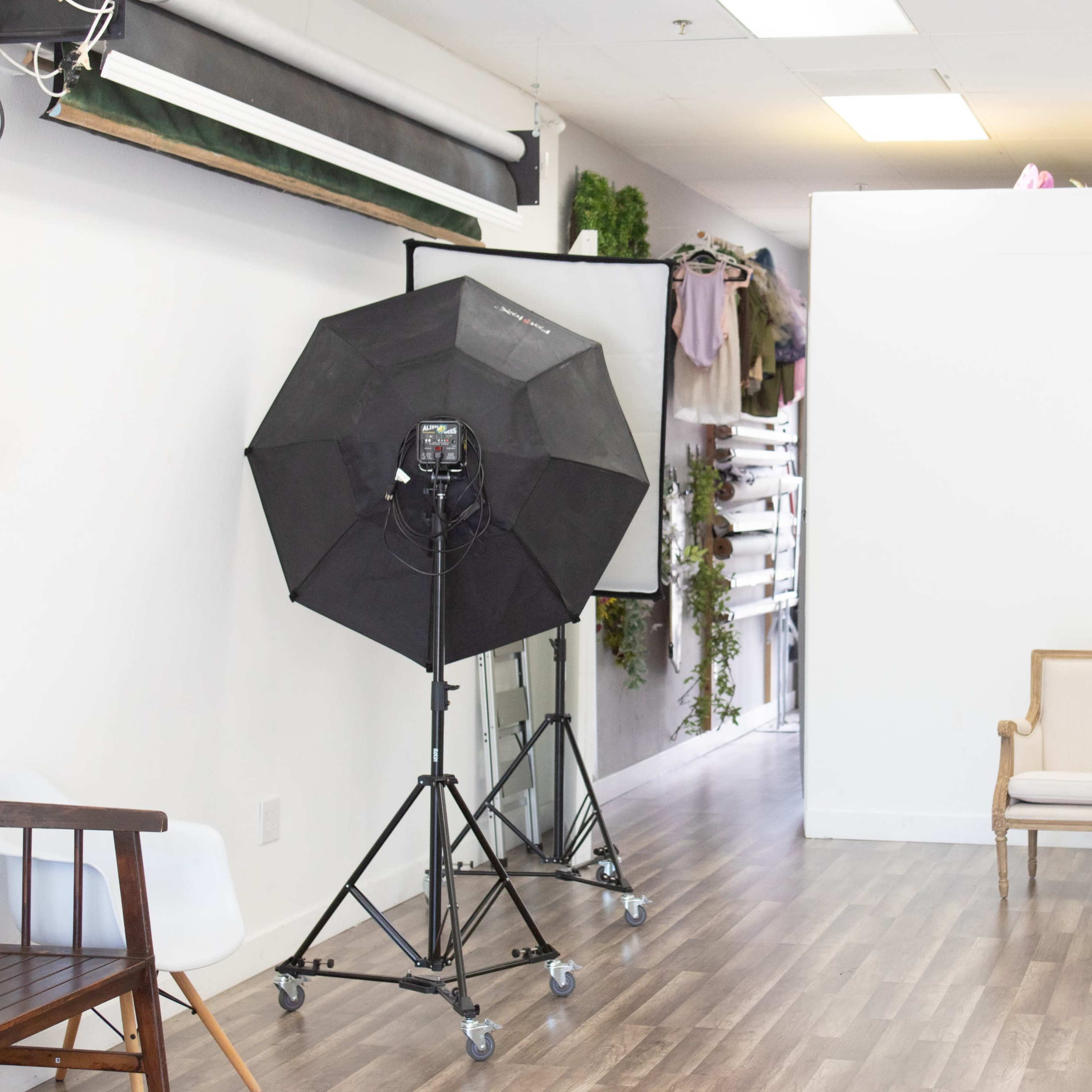 A photography studio featuring a black softbox light on a stand, a wooden bench, and a white backdrop with various props behind it.