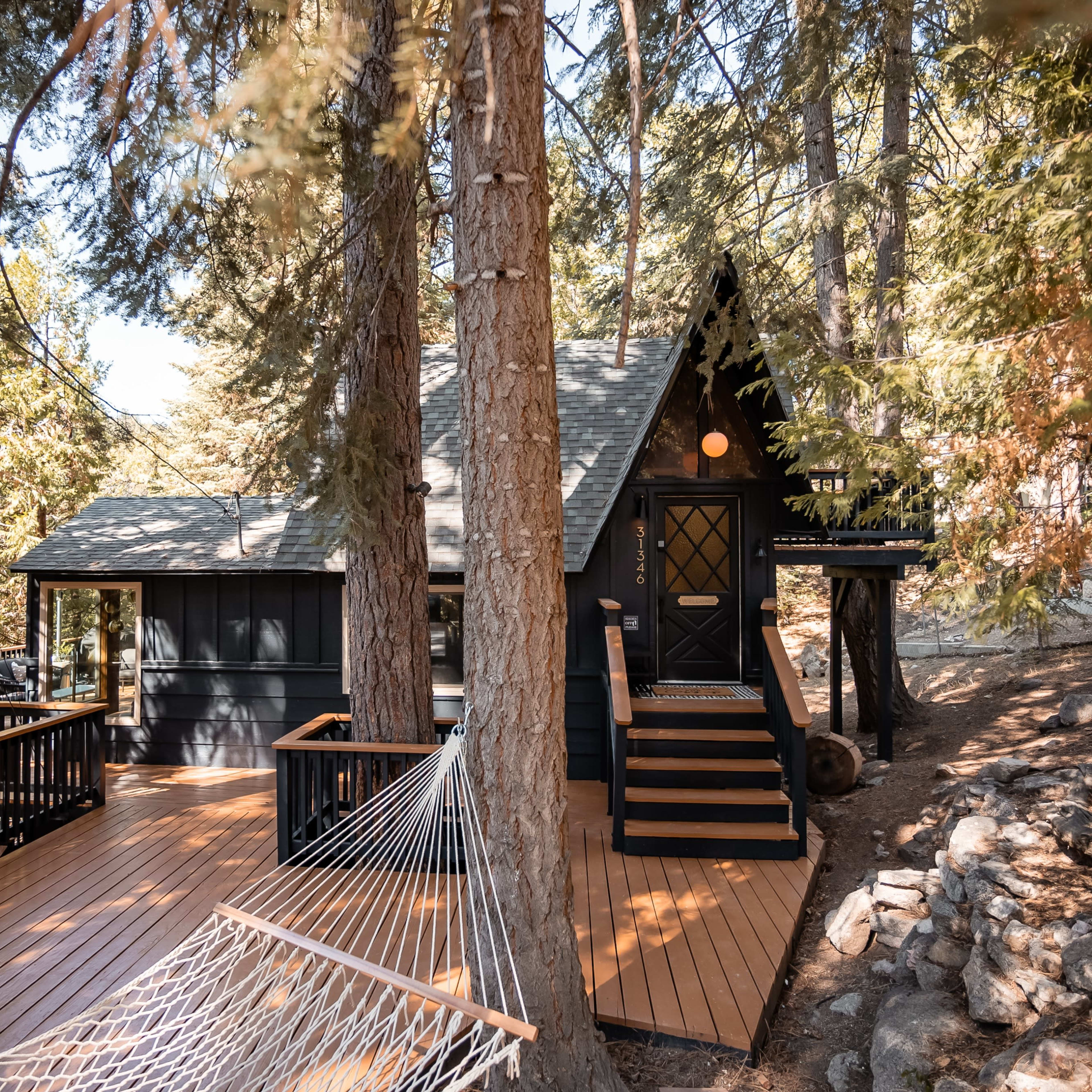 A black chalet-style cabin with a large wooden deck and hammock is surrounded by tall trees in a forested area.