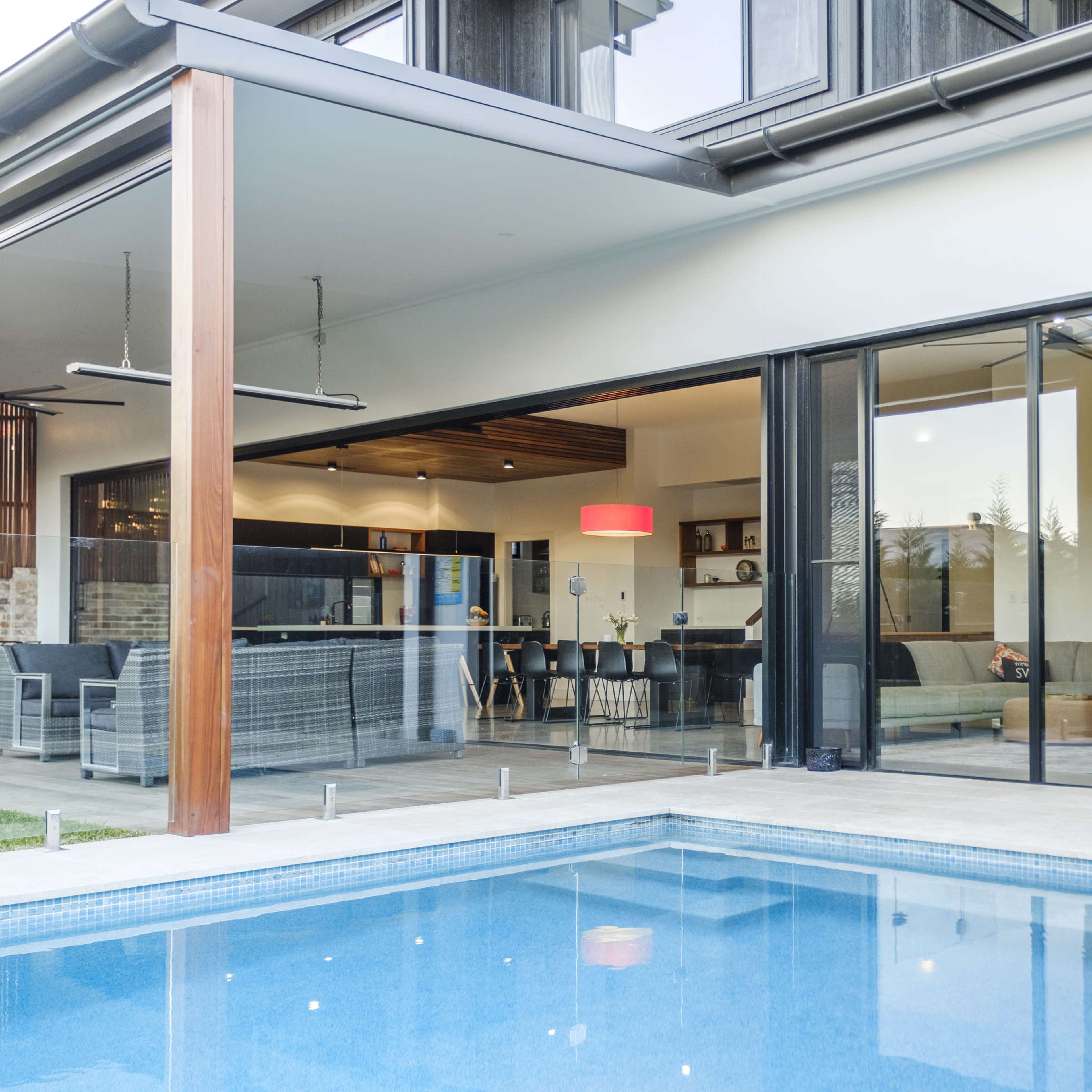 A modern house with large glass doors opens to a pool area, featuring a spacious living room and kitchen visible inside.