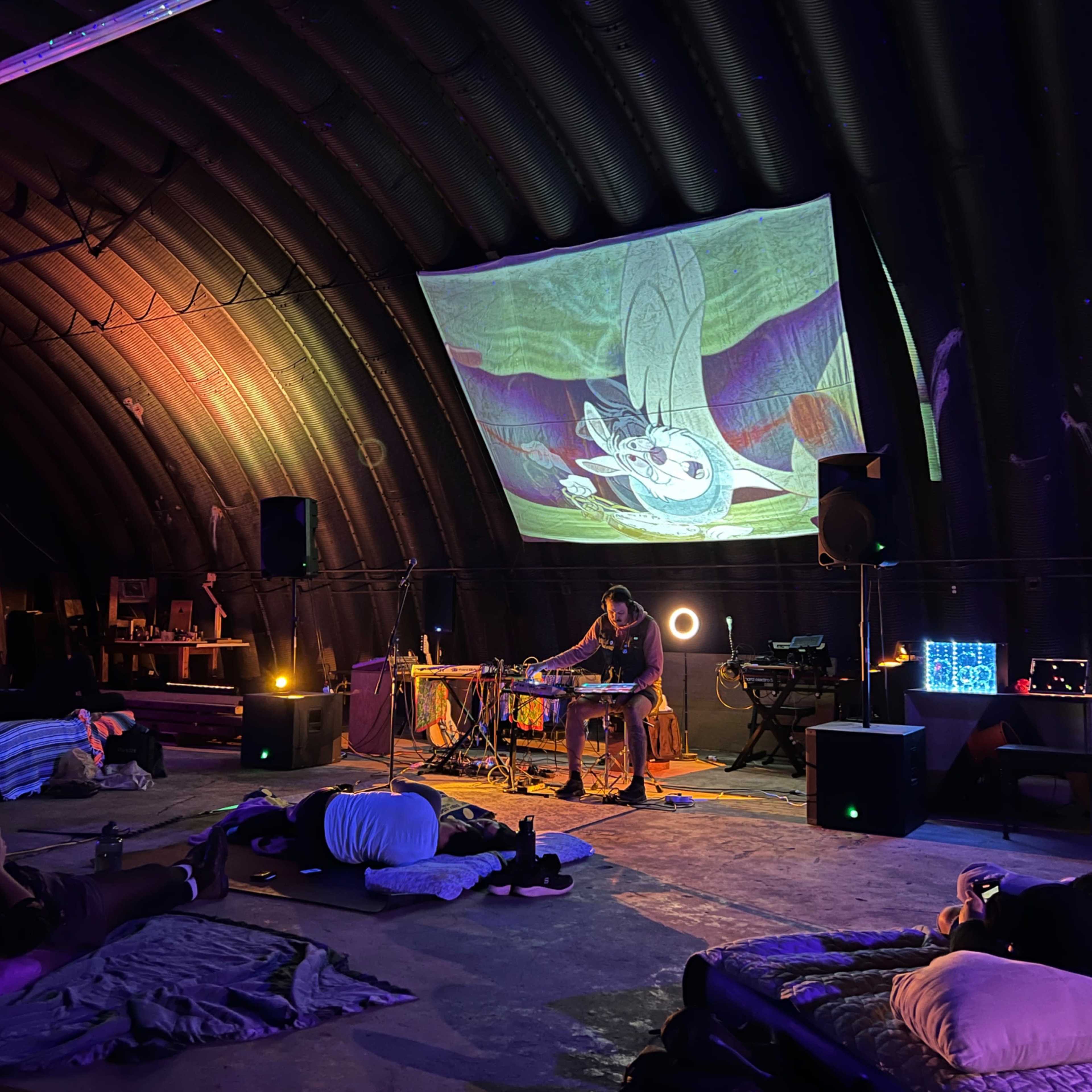 A musician performs on a small stage with a projector display in a dimly lit, spacious indoor venue where people relax on floor cushions.