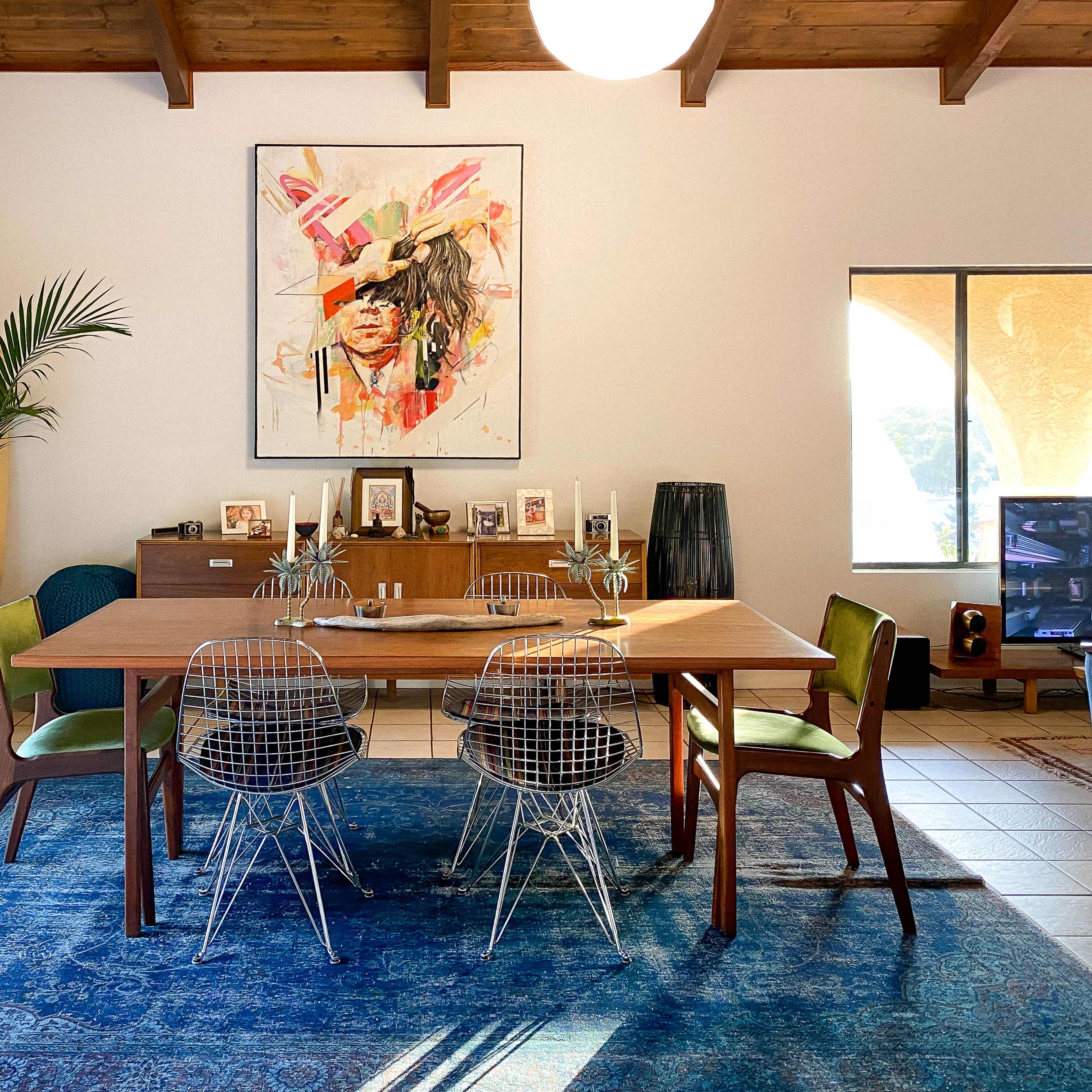 A modern dining space featuring a wooden table surrounded by wire chairs, a large artwork on the wall, and decorative elements including plants and a surfboard.