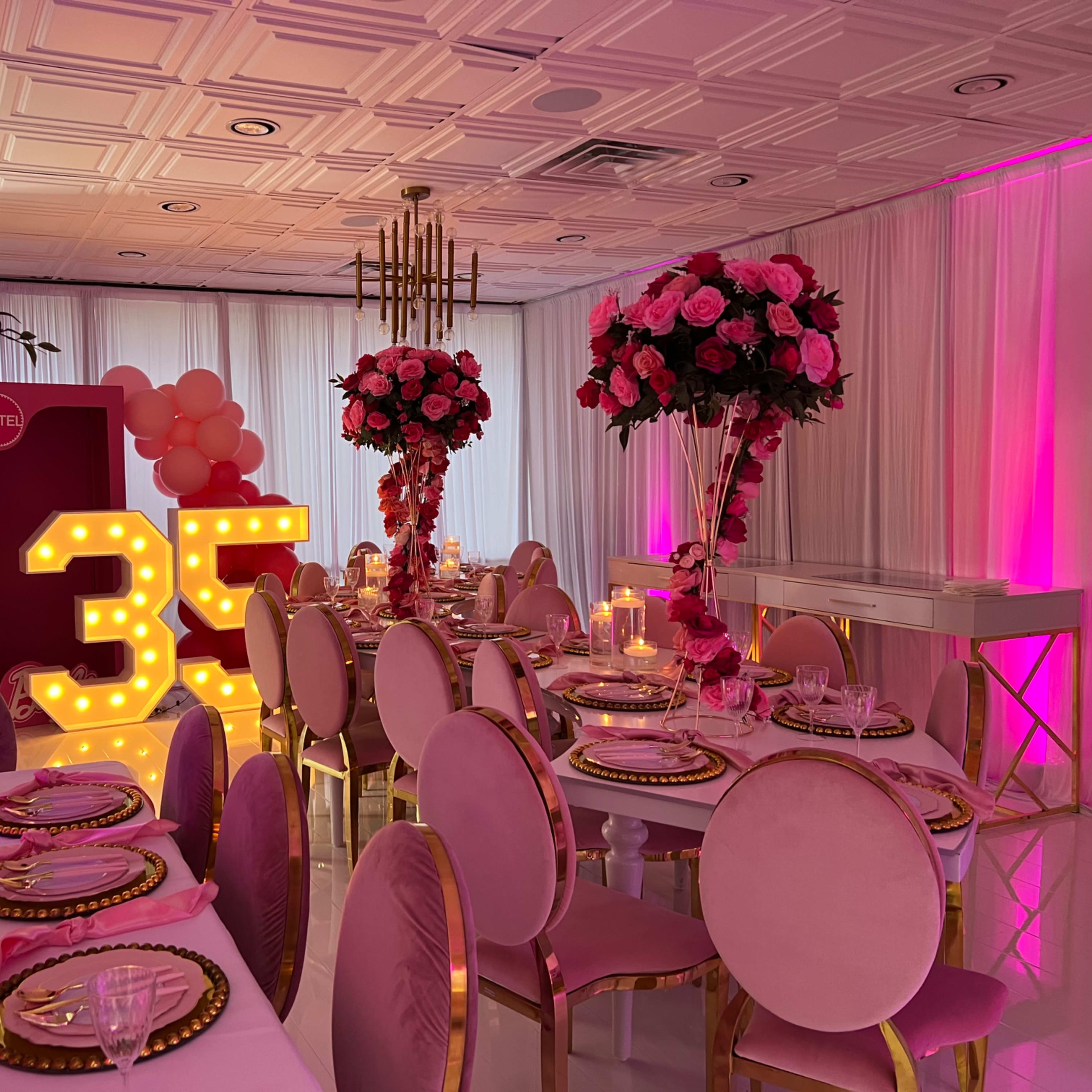 A elegantly decorated banquet hall features large floral centerpieces, a "35" marquee illuminated in bright lights, and tables set with fine dining arrangements under soft pink and white lighting.
