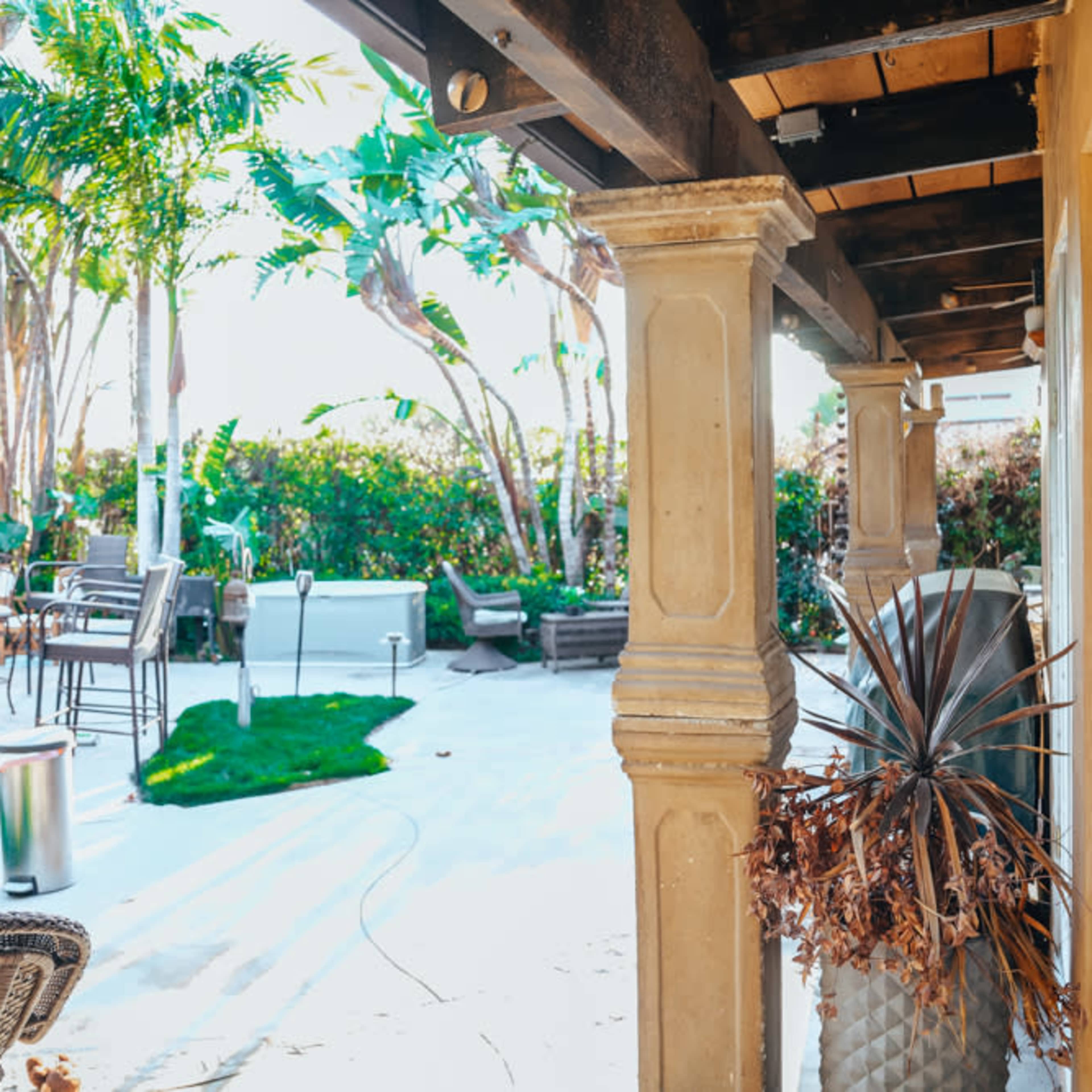A patio area features outdoor furniture, potted plants, and a stone column under a wooden roof, surrounded by palm trees and greenery.