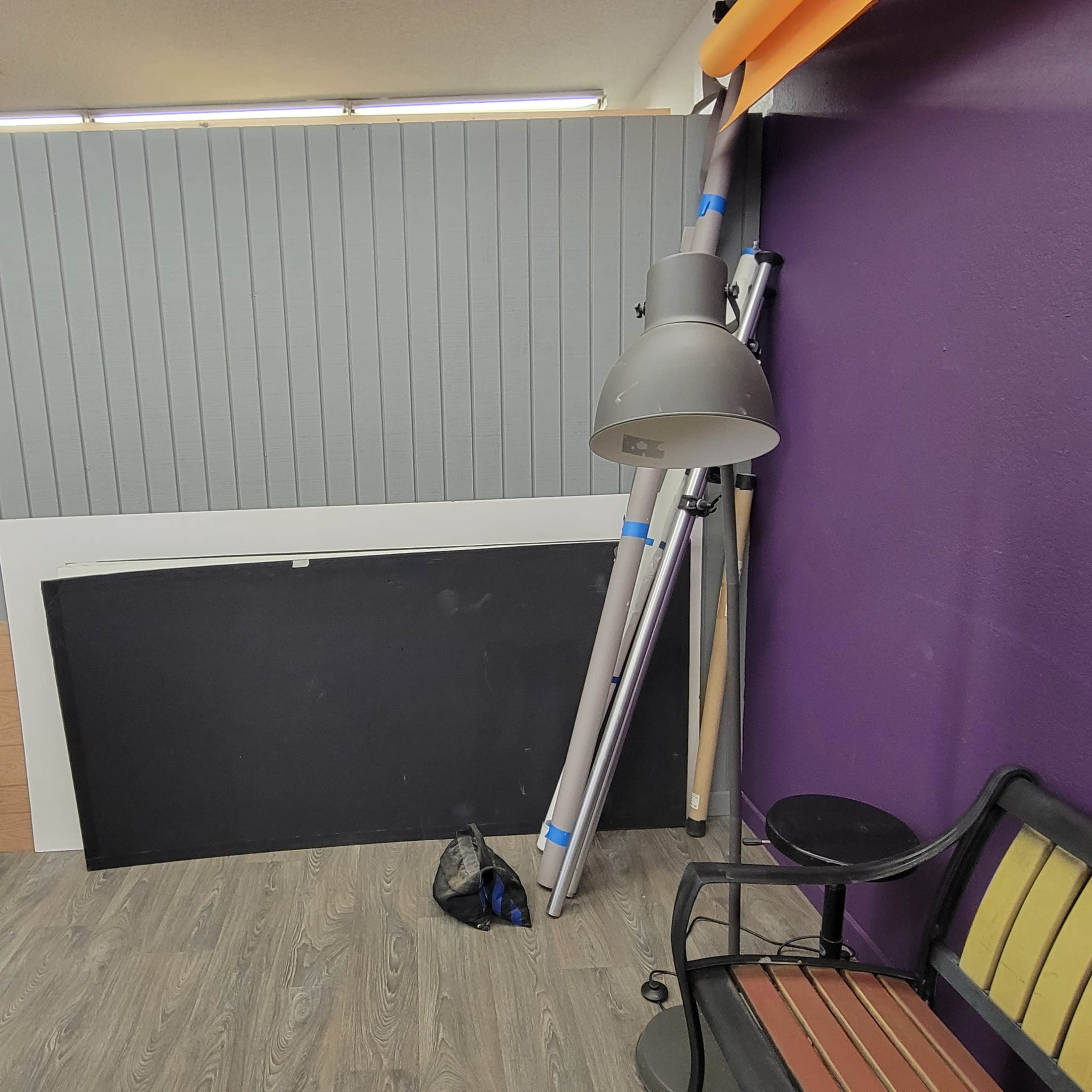 A room setup with a purple wall, a gray panel, photography equipment including softboxes, a tripod, and a floor lamp beside a seating area.
