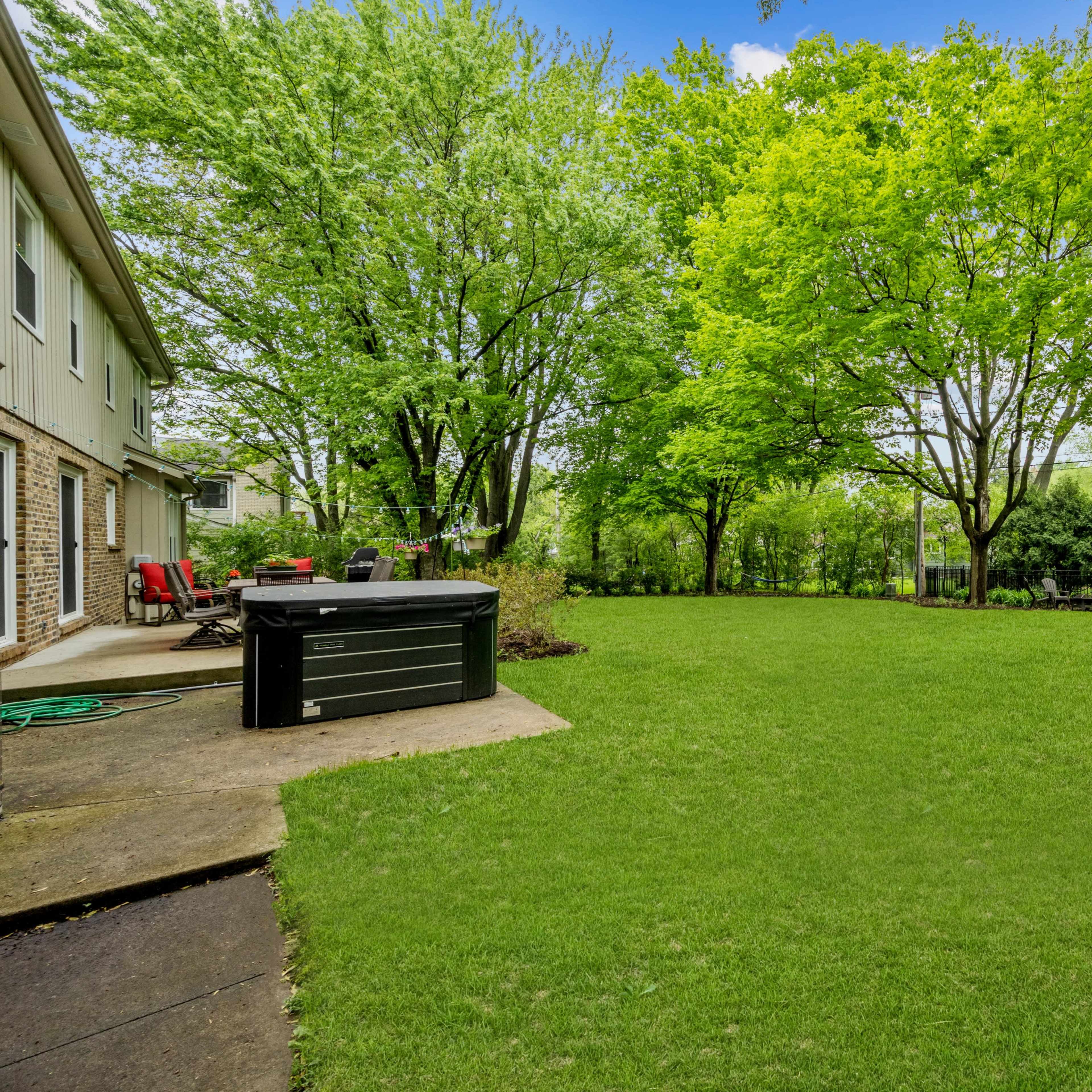 A spacious backyard with well-maintained grass, surrounded by trees, and features a hot tub on a patio.
