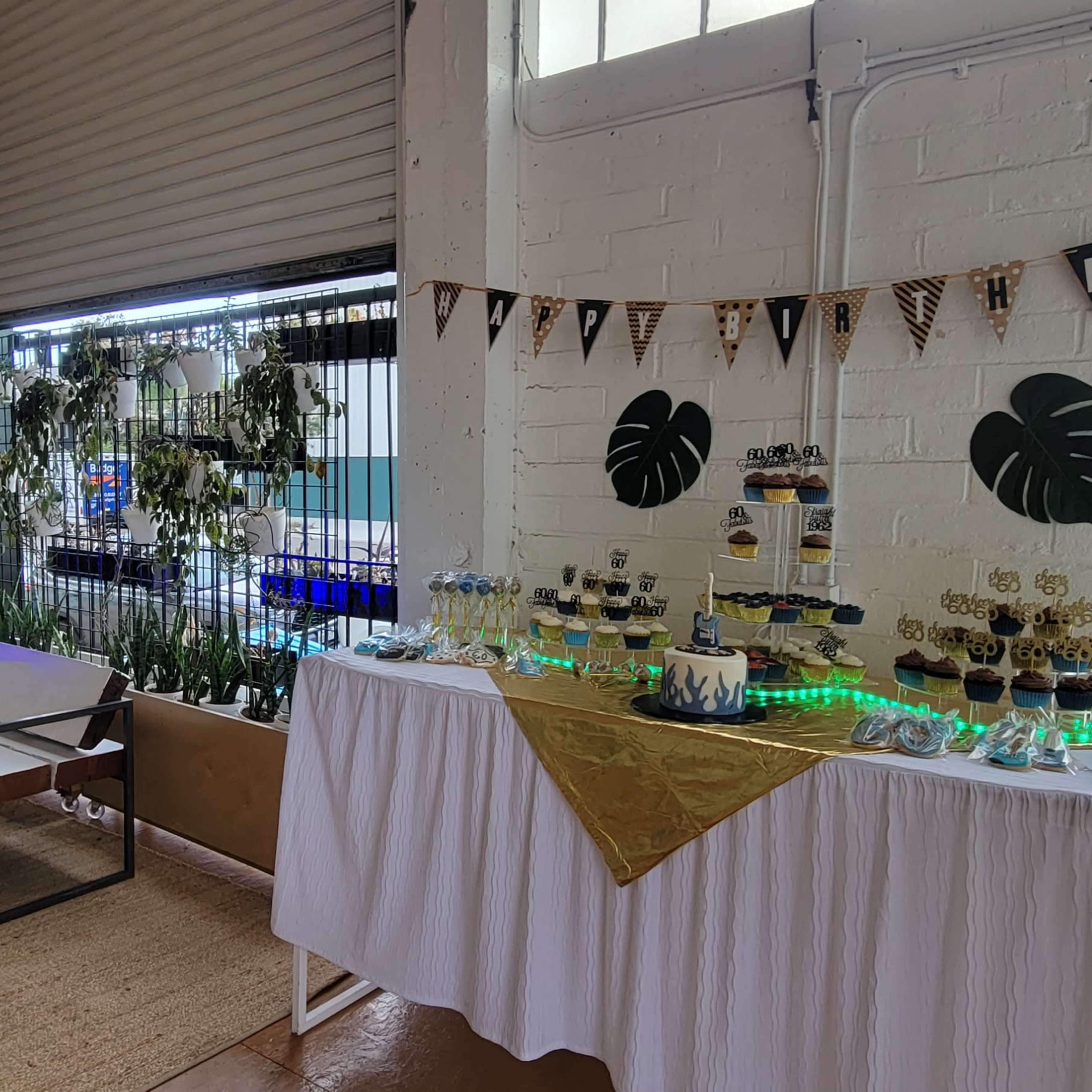 A decorated indoor space featuring a table with desserts and plants, along with seating arrangements and large windows.