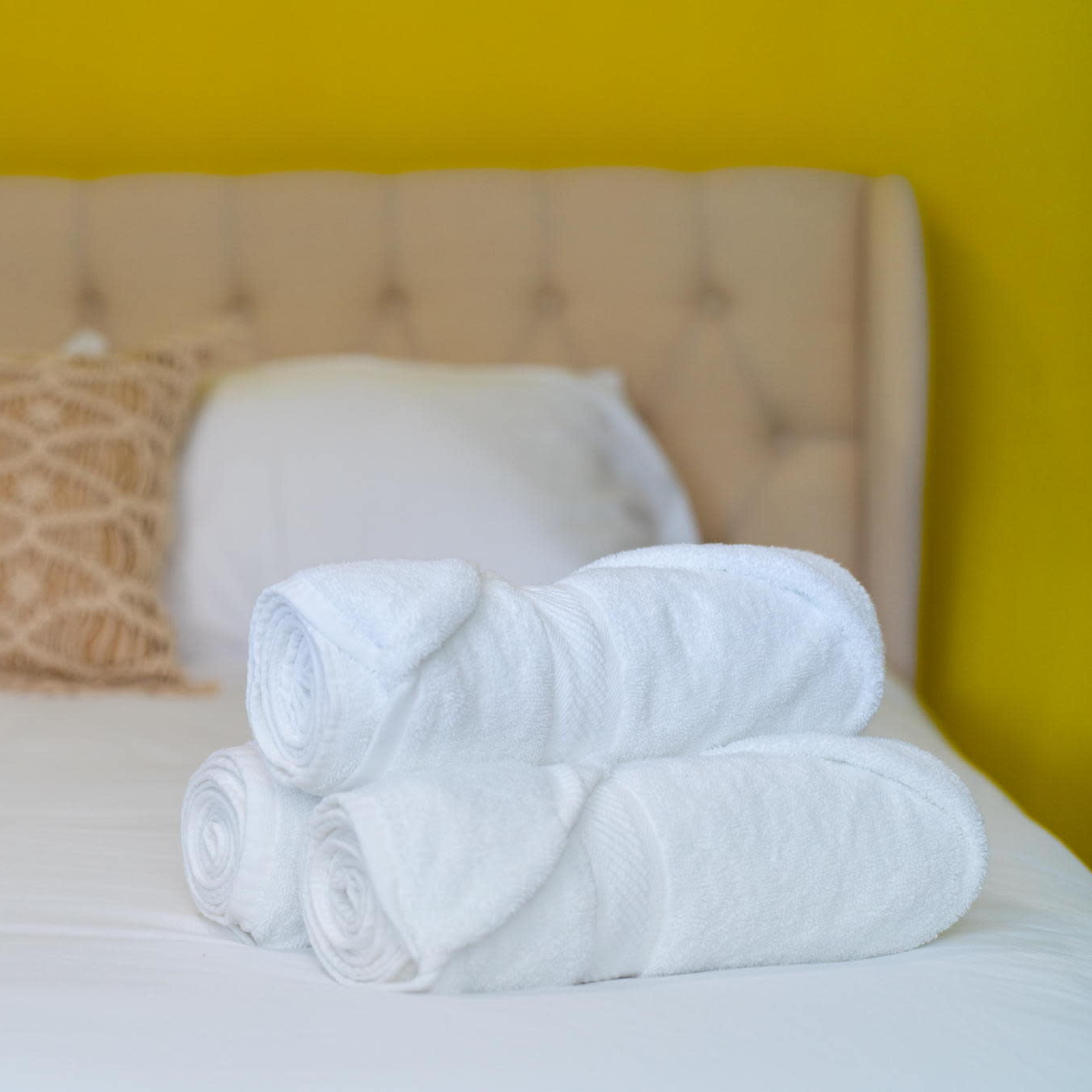 A neatly arranged stack of white towels is placed on a bed with a tufted headboard and a decorative pillow against a yellow wall.
