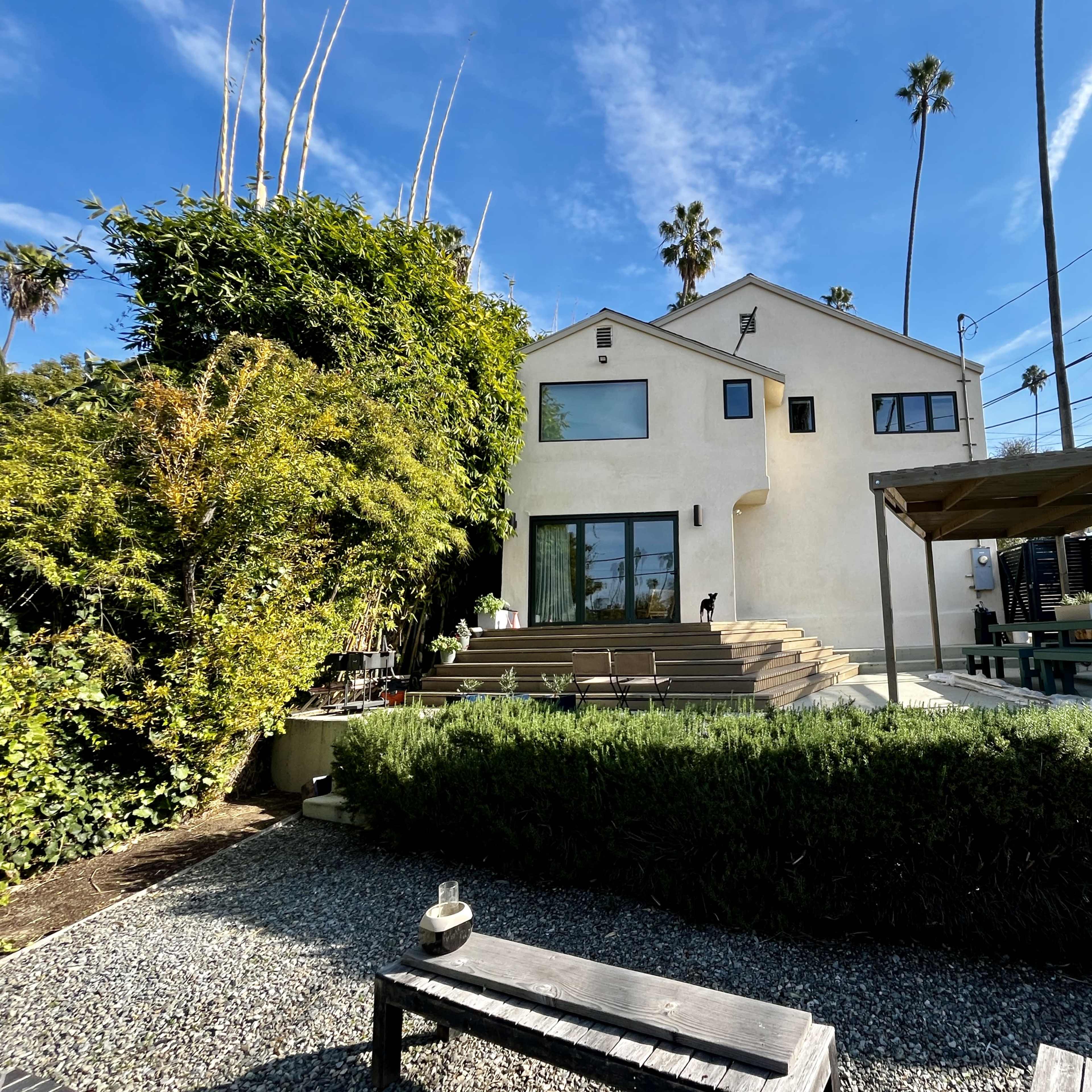 A modern house with a landscaped garden, featuring wooden steps and a patio area, surrounded by palm trees and power lines.