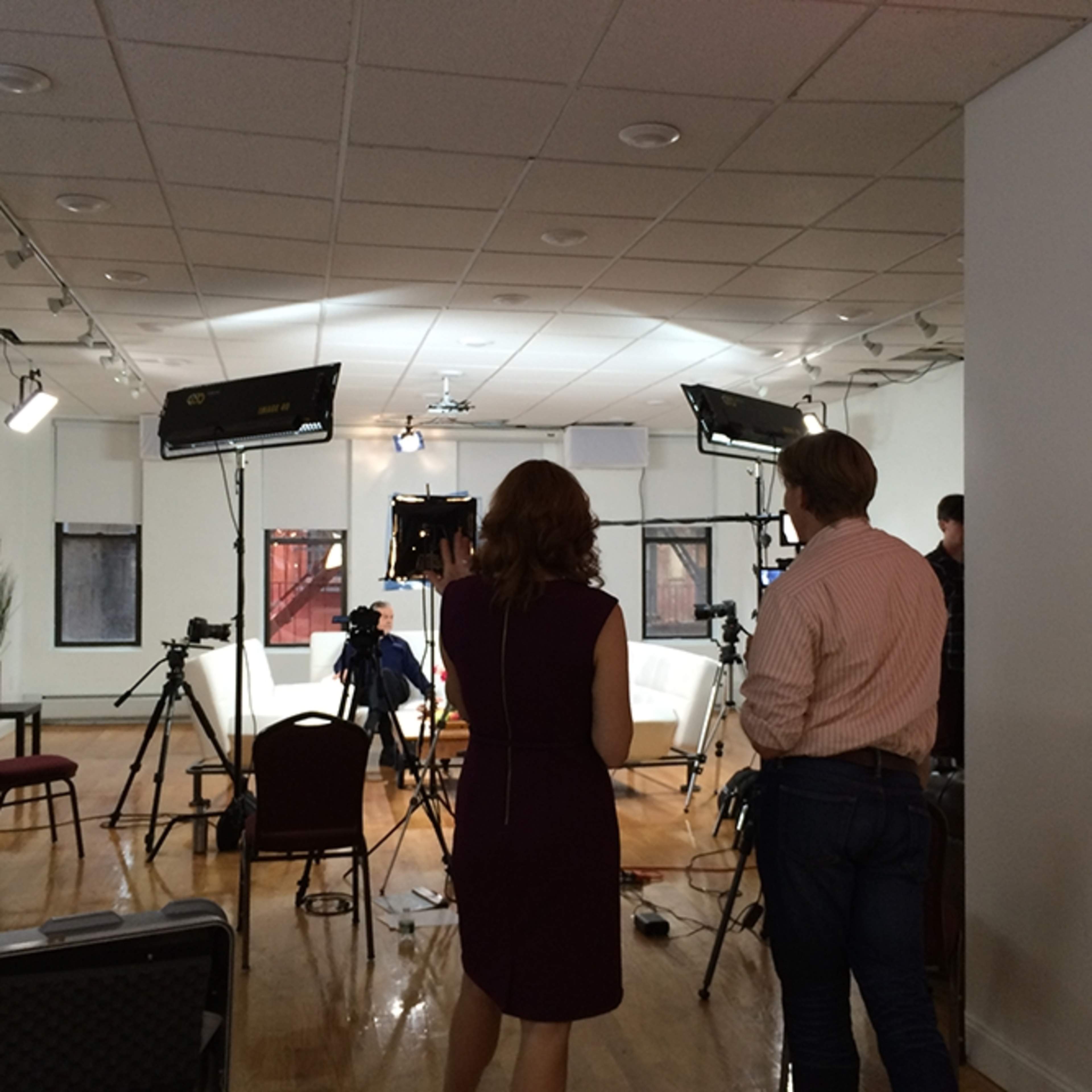 A film set is arranged in a room with multiple cameras and lighting equipment, where two people stand discussing the setup.