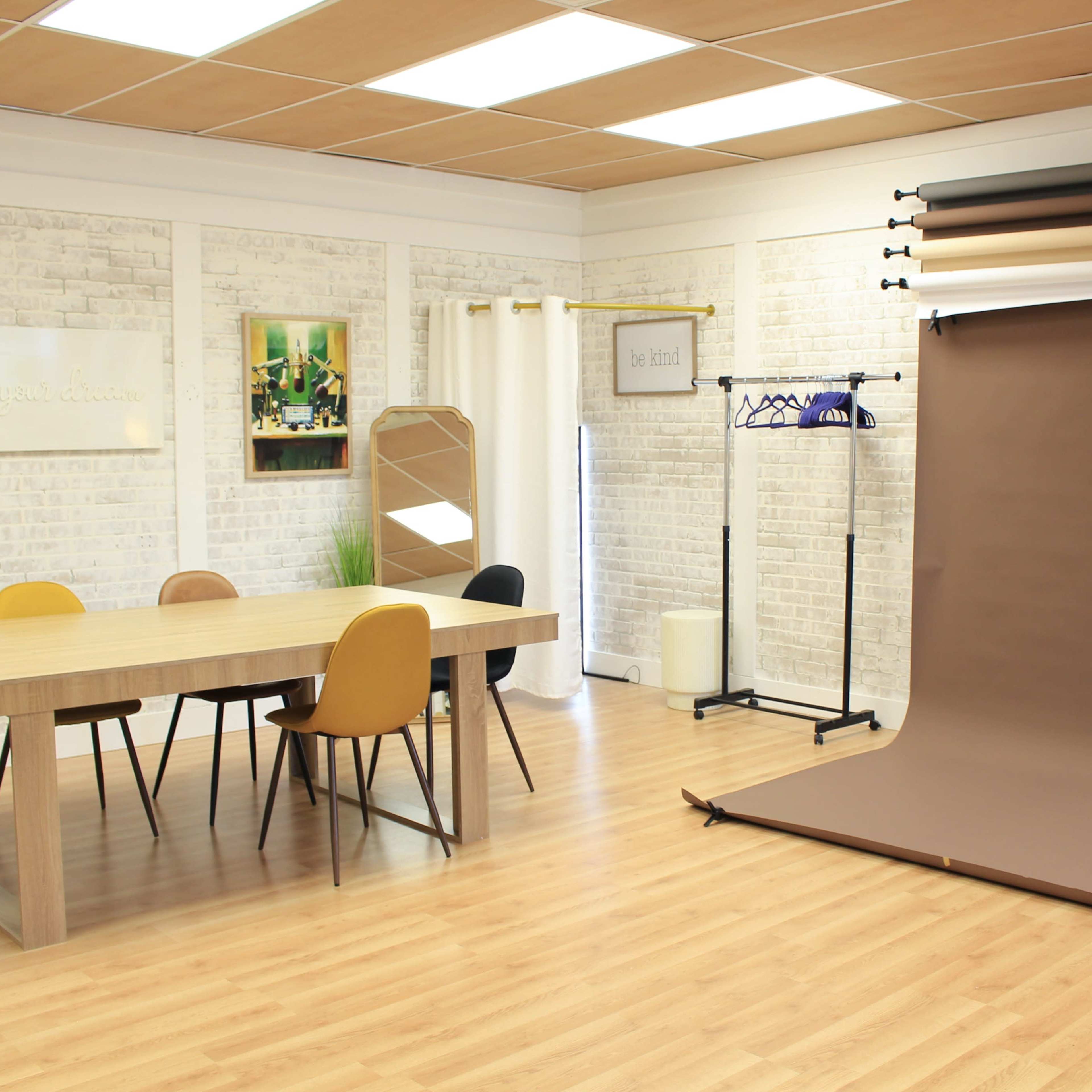 A bright, minimalist studio space with a wooden table, mixed-color chairs, and a backdrop stand featuring various fabric rolls.