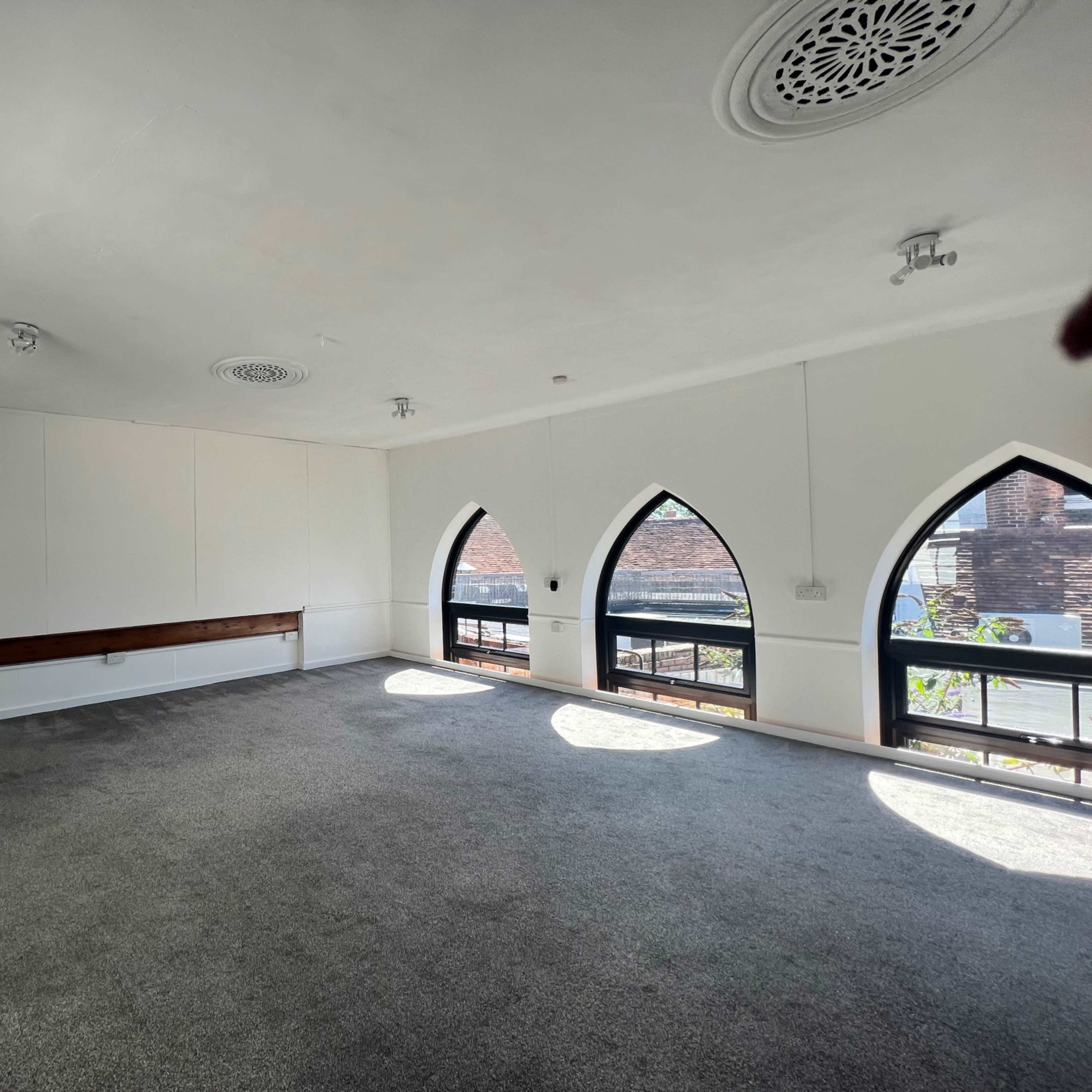 A spacious room with carpeted floors and large arched windows allowing light to enter.