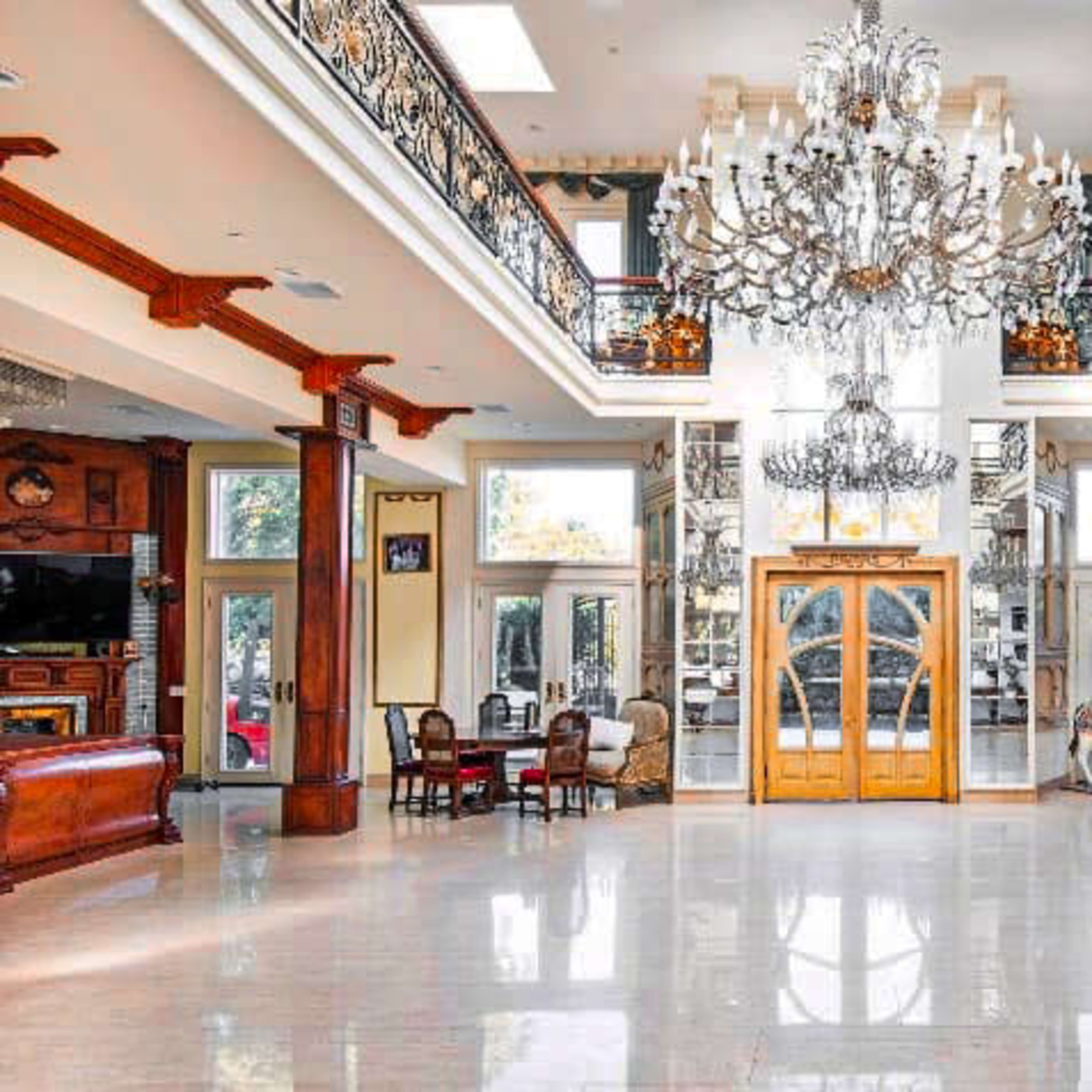 An expansive foyer with a high ceiling, featuring a grand chandelier, elegant wooden doors, and a polished marble floor.