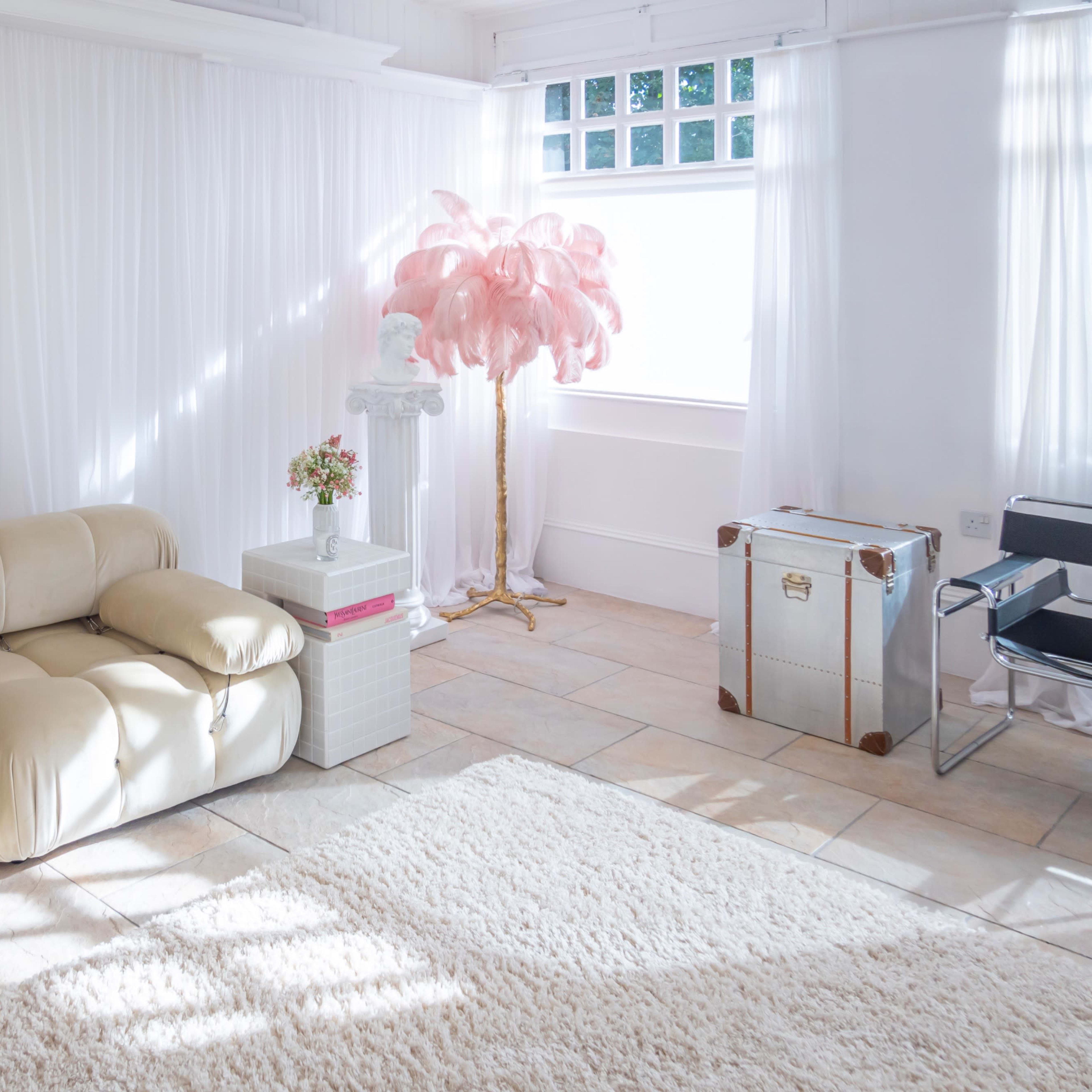 A bright, minimalist living space with a cream-colored sofa, a small table topped with flowers, a pink decorative tree, and a metallic trunk.