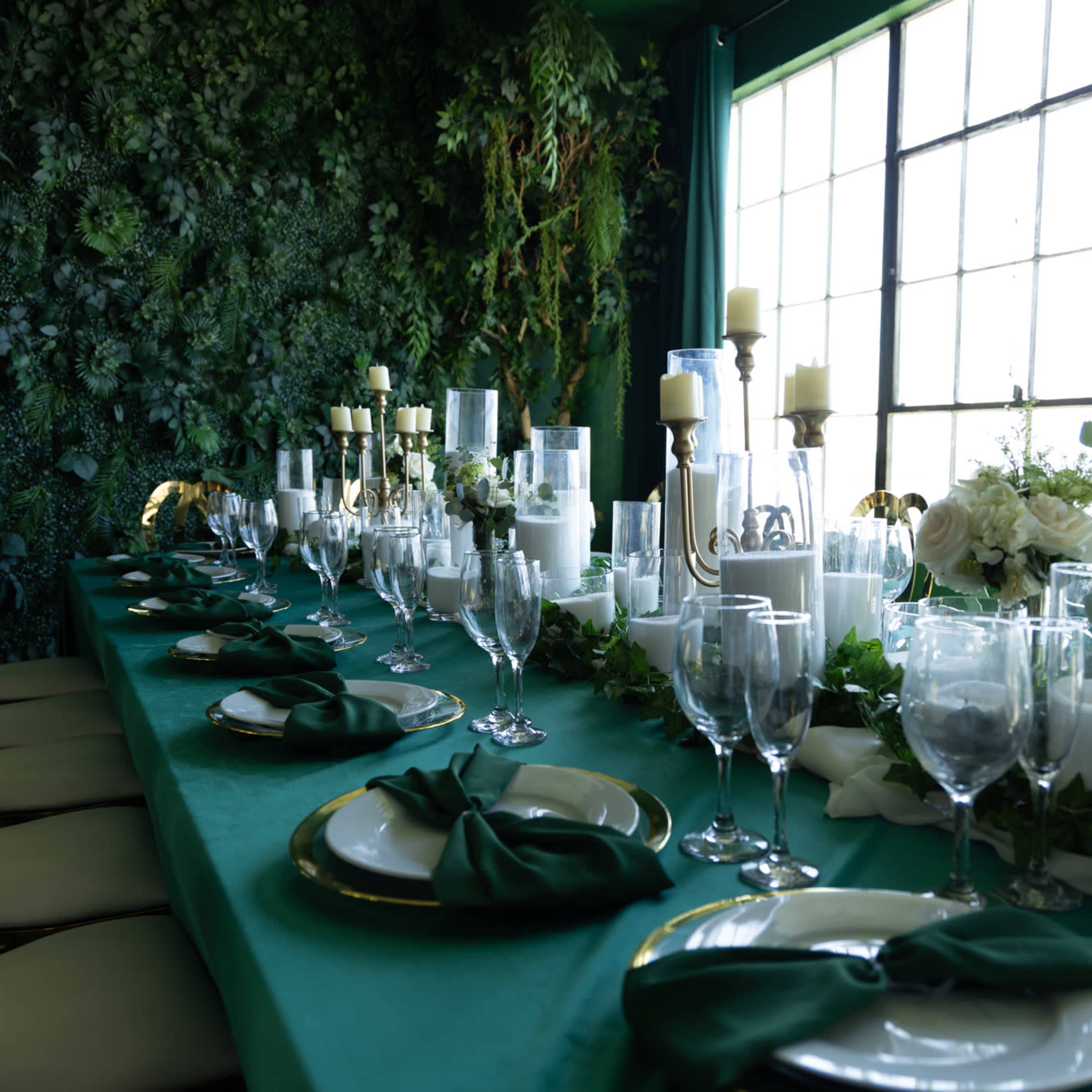 A long dining table is set with white candles, glassware, and green napkins against a backdrop of lush greenery.