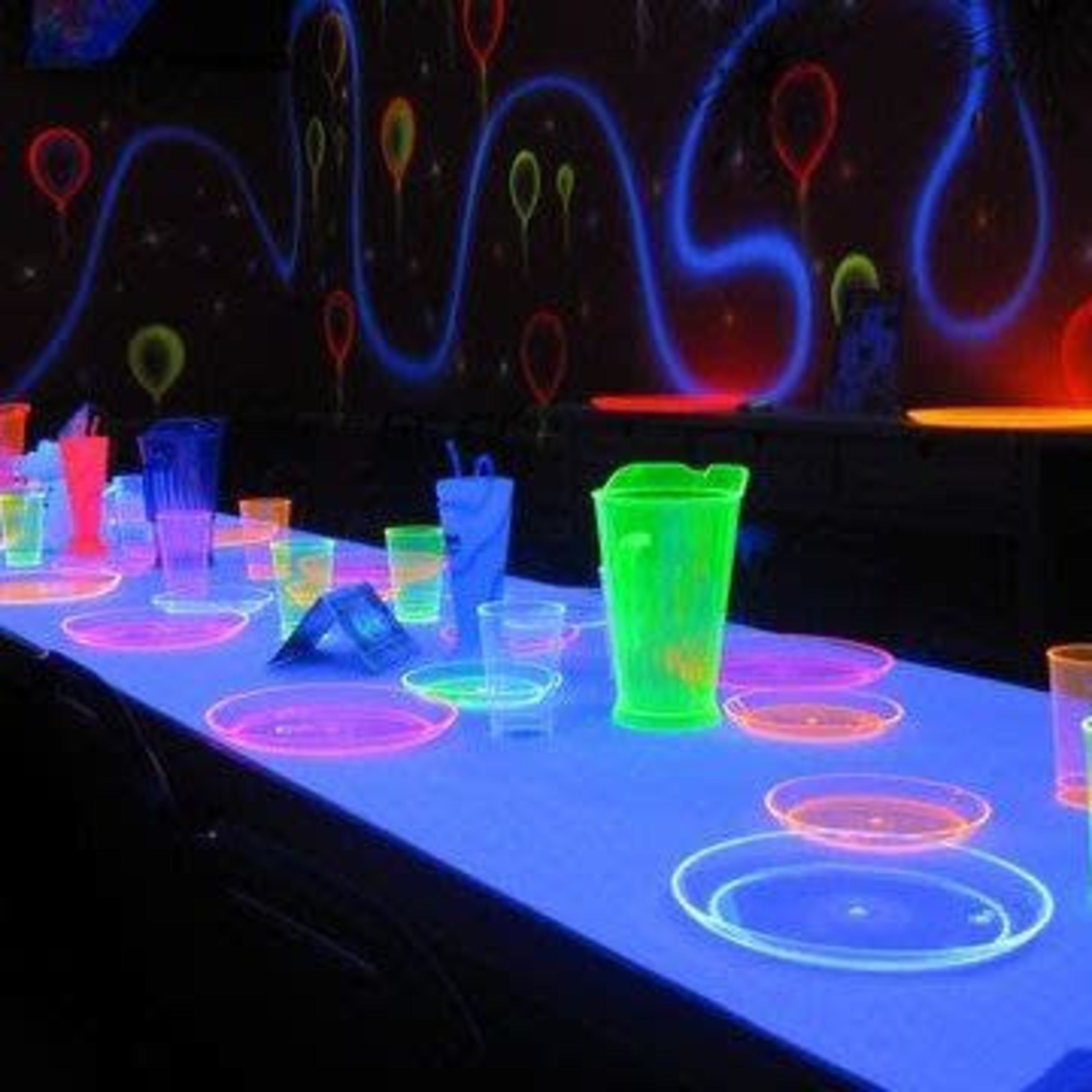 A long table is set with glowing neon dinnerware and utensils under a blacklight.