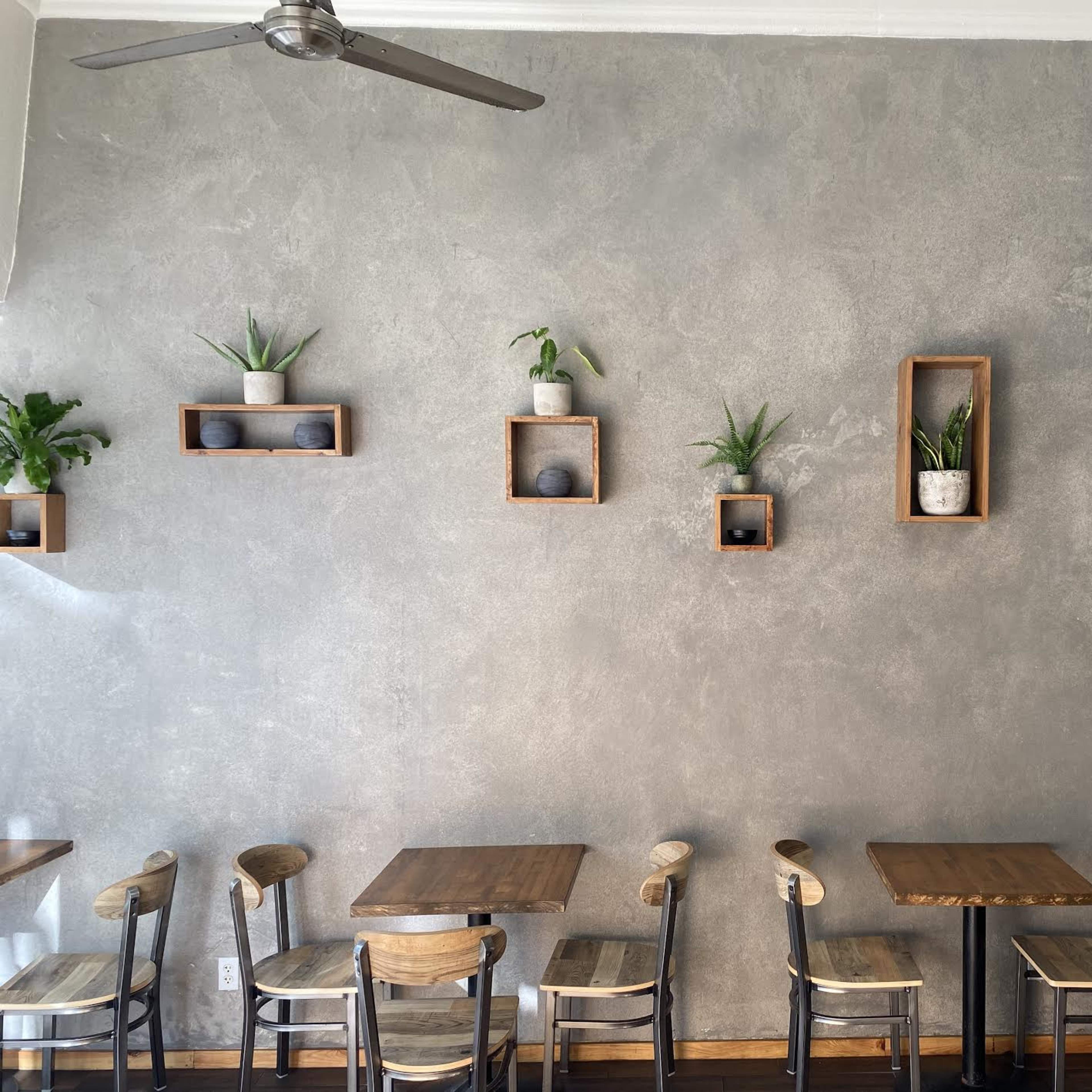 A café interior featuring wooden tables and chairs, with a textured gray wall adorned with floating shelves displaying potted plants.