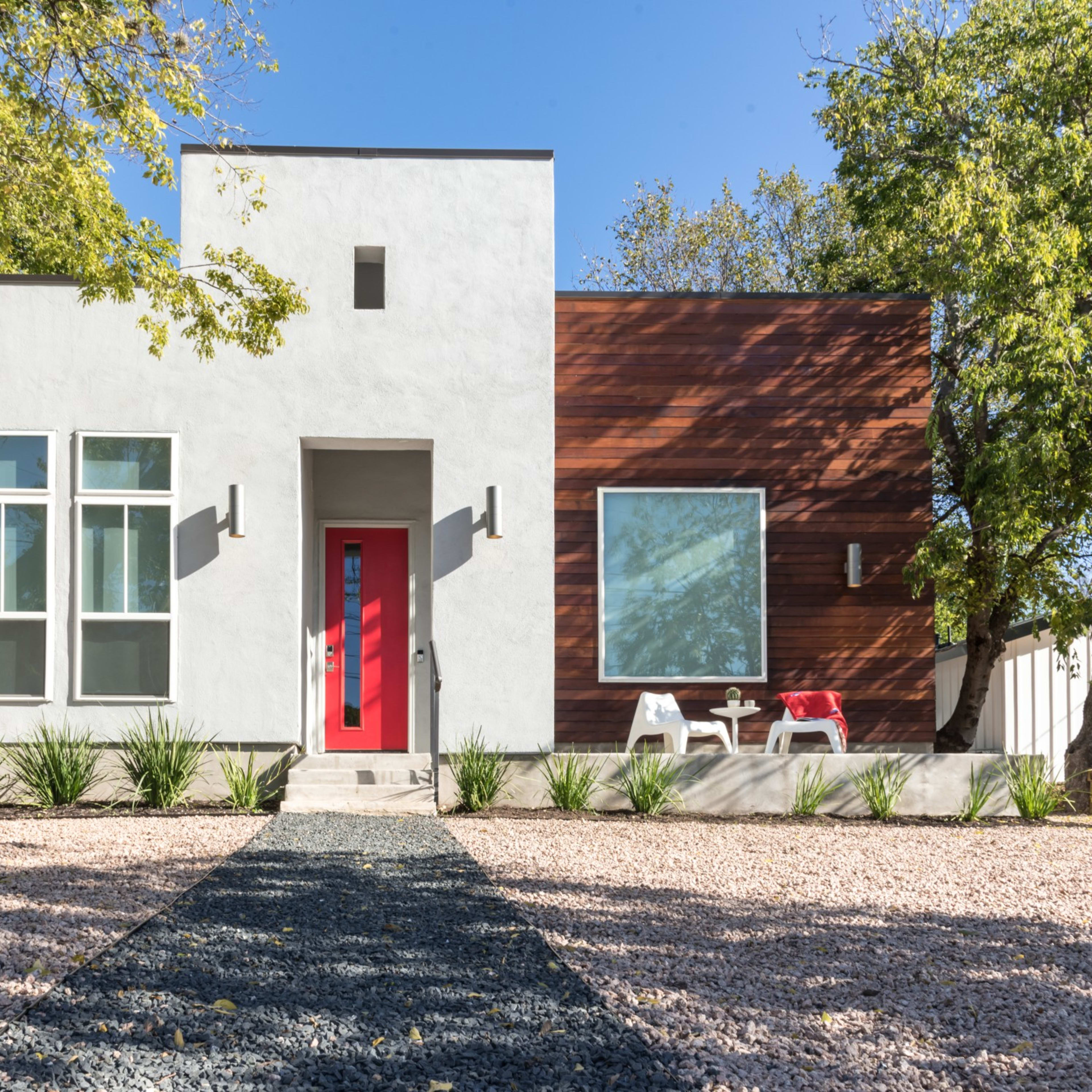 A modern house features a white exterior with wooden accents, a bright red door, and landscaped gravel pathways.