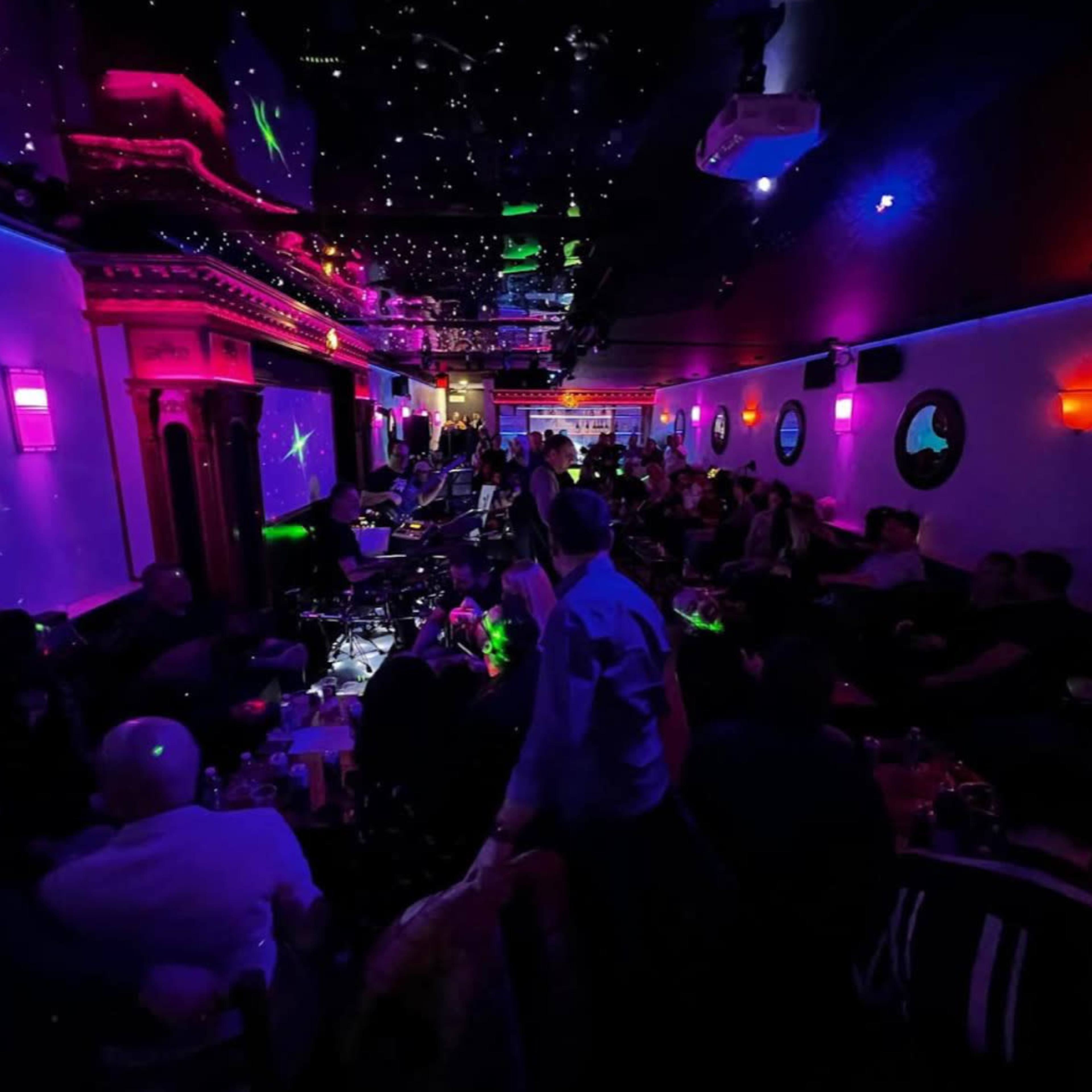 A crowded nightclub features colorful lighting and a live band performing on stage.