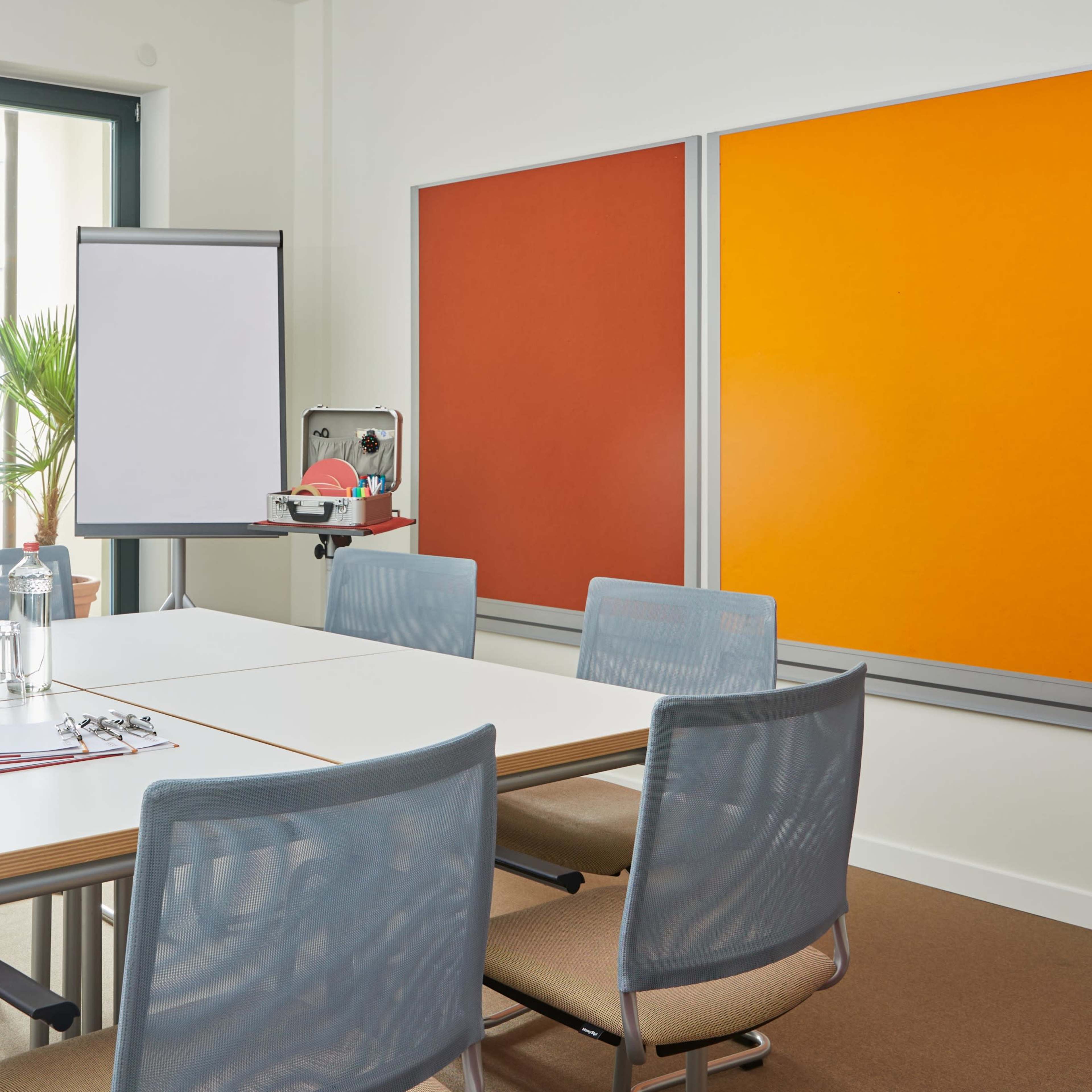 A modern conference room with a large table, several chairs, a flip chart, and large windows allowing natural light.