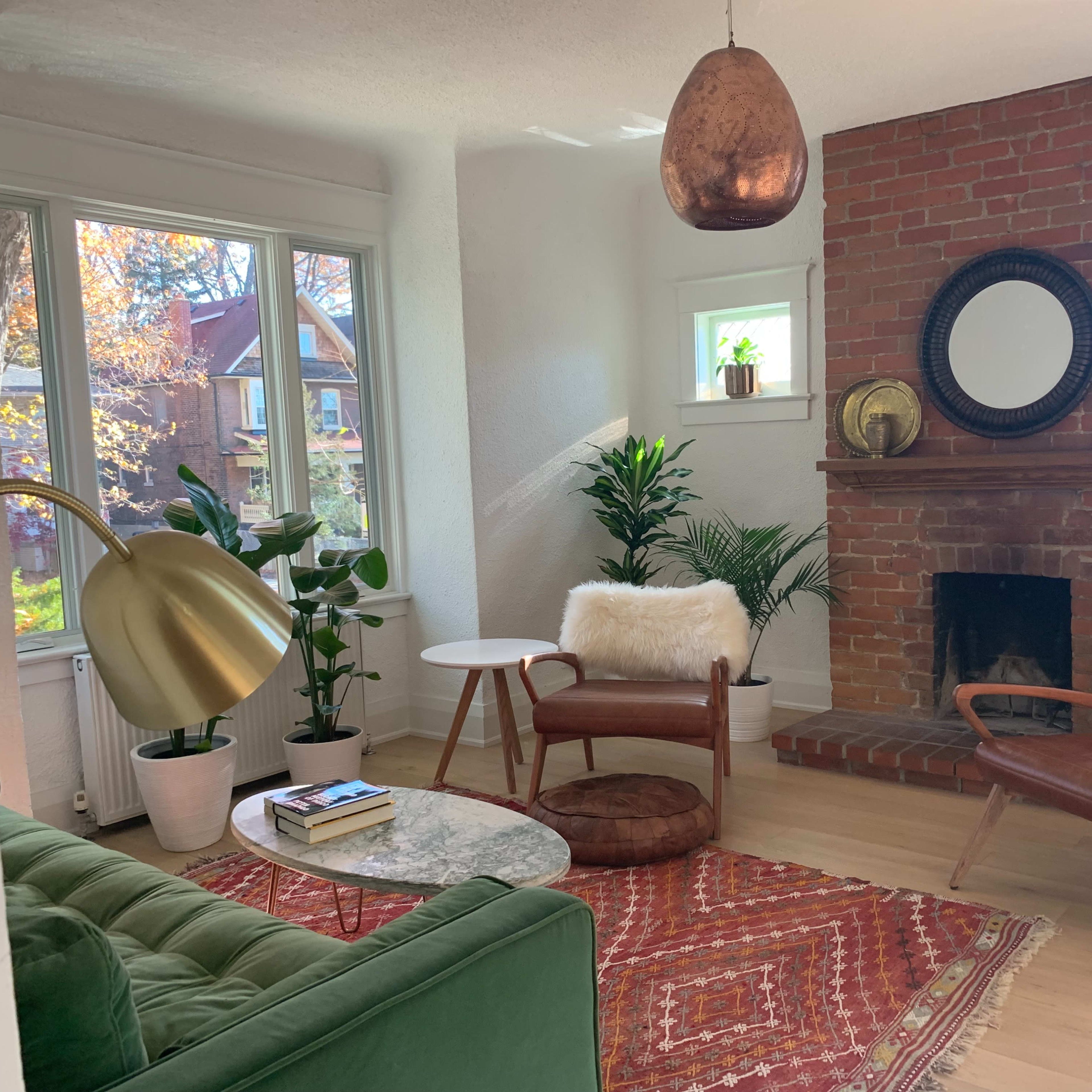 A cozy living room features a green couch, a round marble coffee table, wooden chairs with fur cushions, and large plants near a brick fireplace.