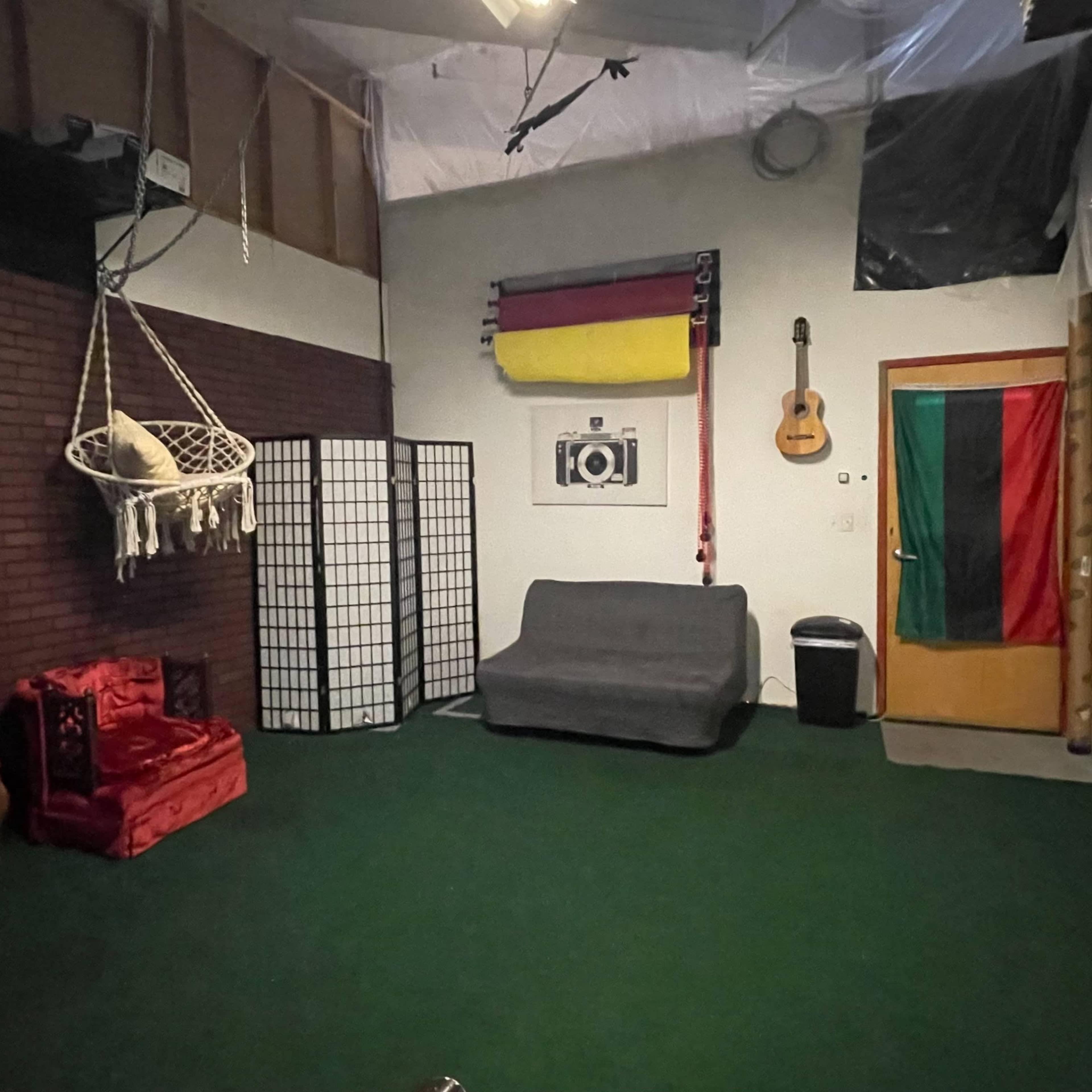 A room with green carpet, a gray couch, a hanging chair, and a door with flags and a guitar on the wall.