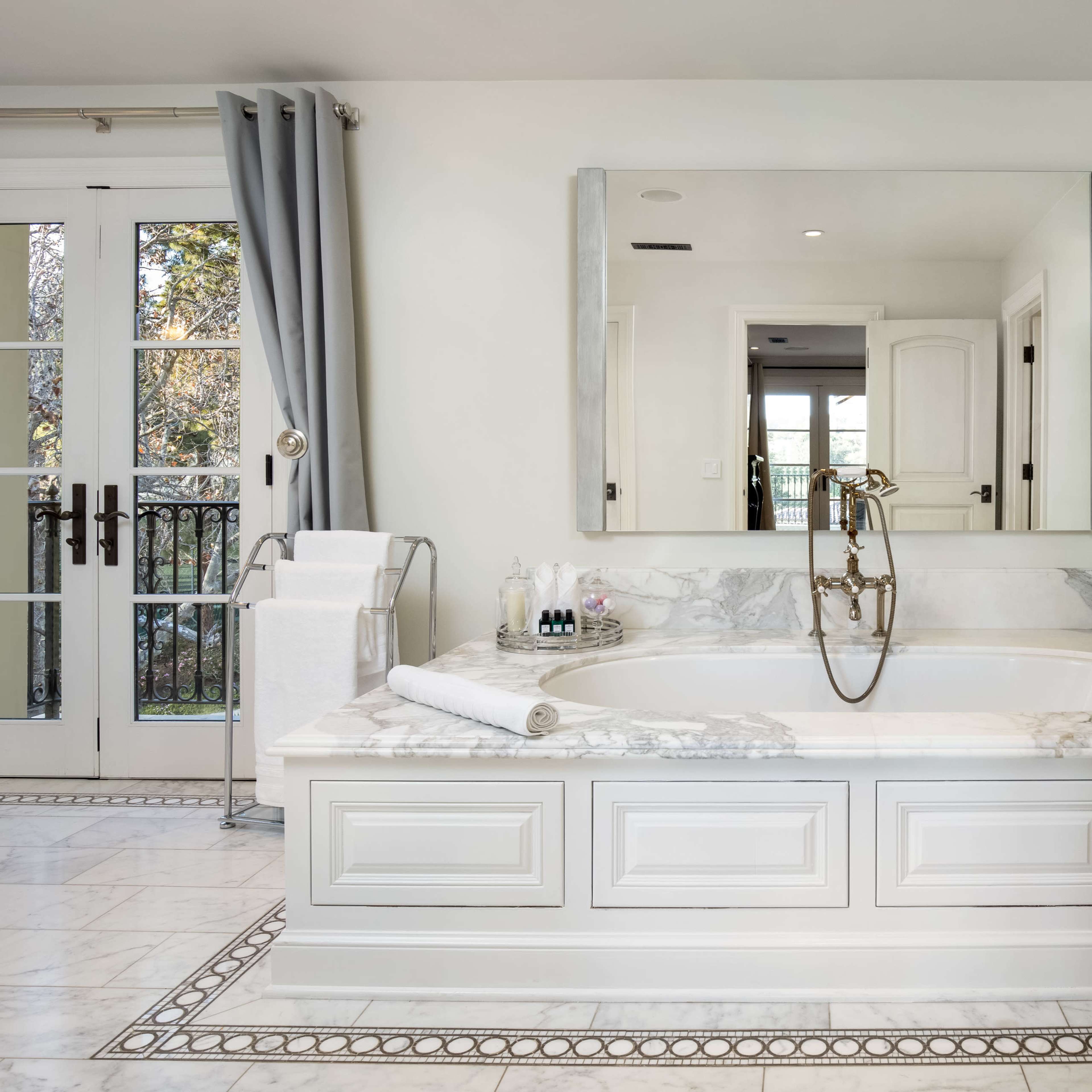 A luxurious marble bathtub is positioned next to a large mirror and ample natural light from French doors.