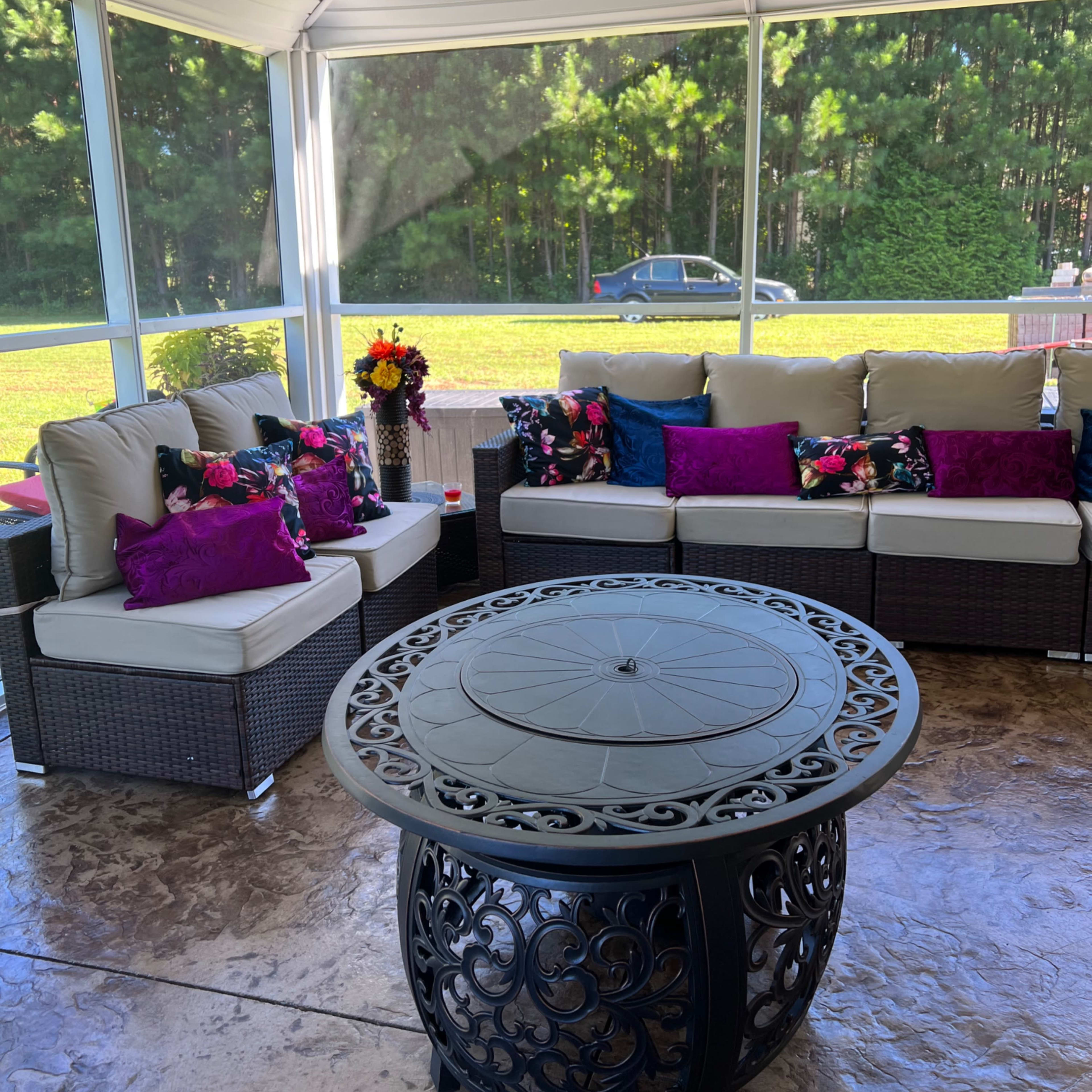 A screened-in porch features a seating area with white cushions and colorful pillows around a circular metal coffee table, surrounded by greenery.