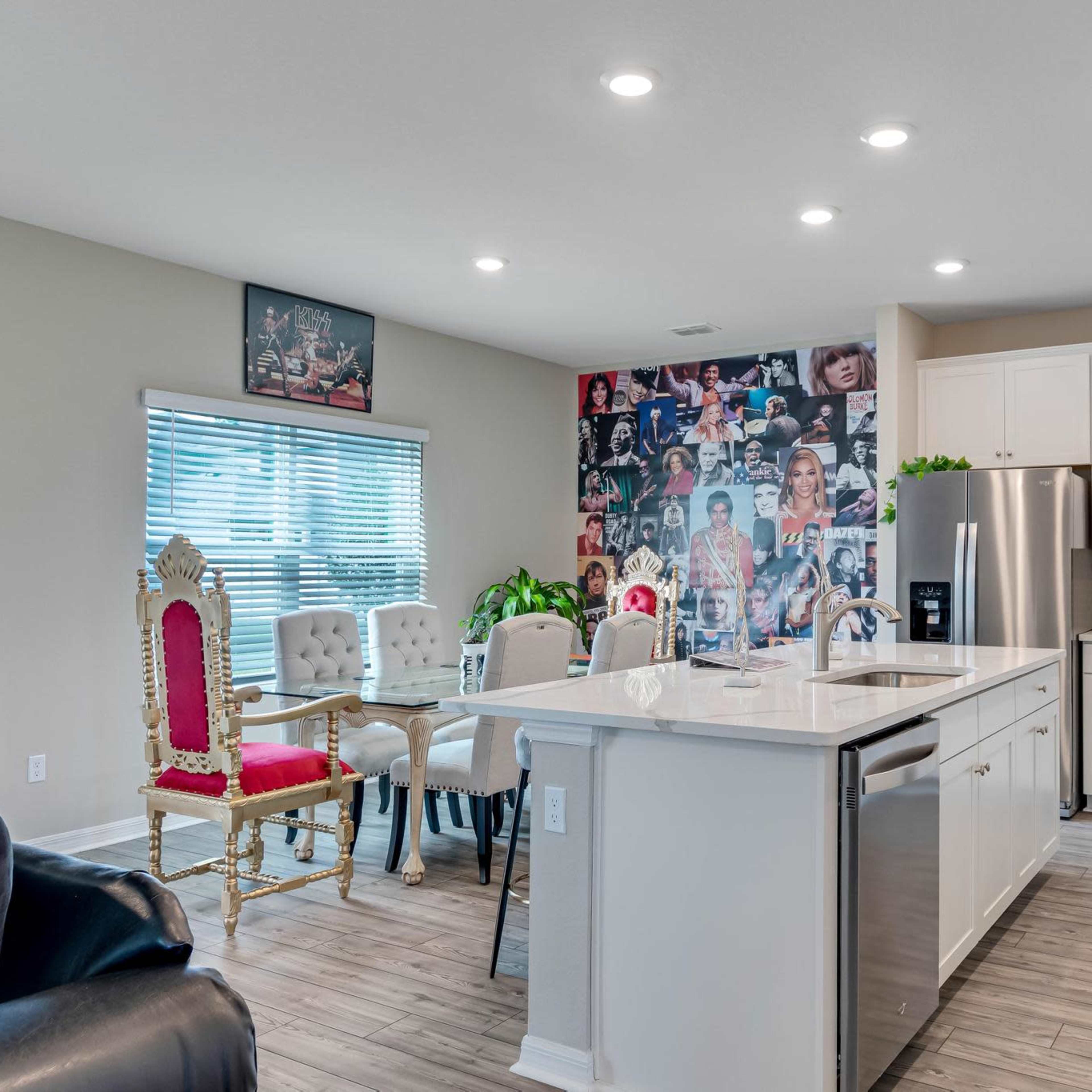 A modern kitchen features white cabinetry, a central island with a sink, and a dining area with upholstered chairs, alongside a wall adorned with a collage of images.