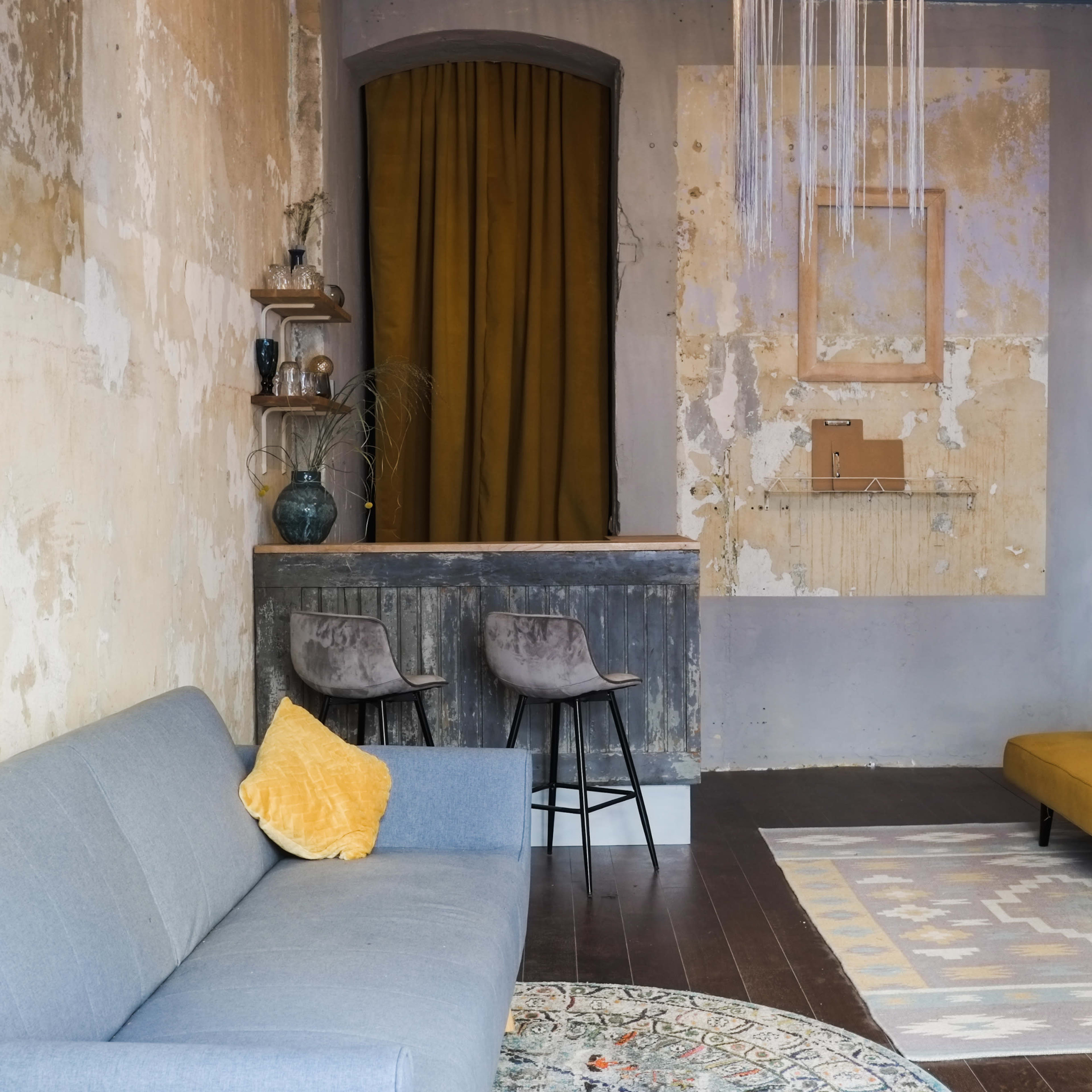 A room with a gray sofa, a yellow seating area, a small bar with two stools, and walls featuring peeling paint and a large circular decoration.