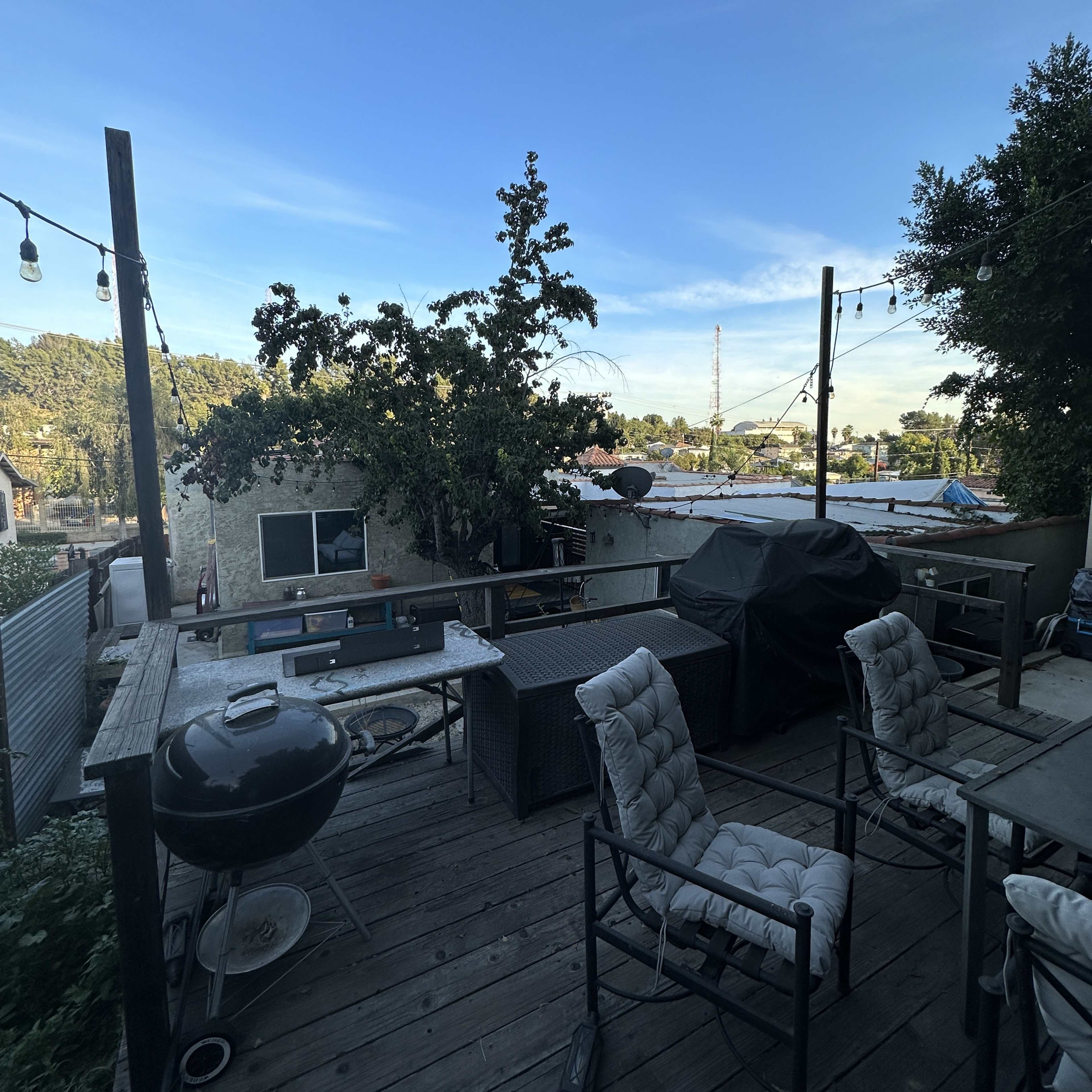A wooden deck with outdoor furniture and a grill overlooks a backdrop of trees and rooftops under a clear blue sky.
