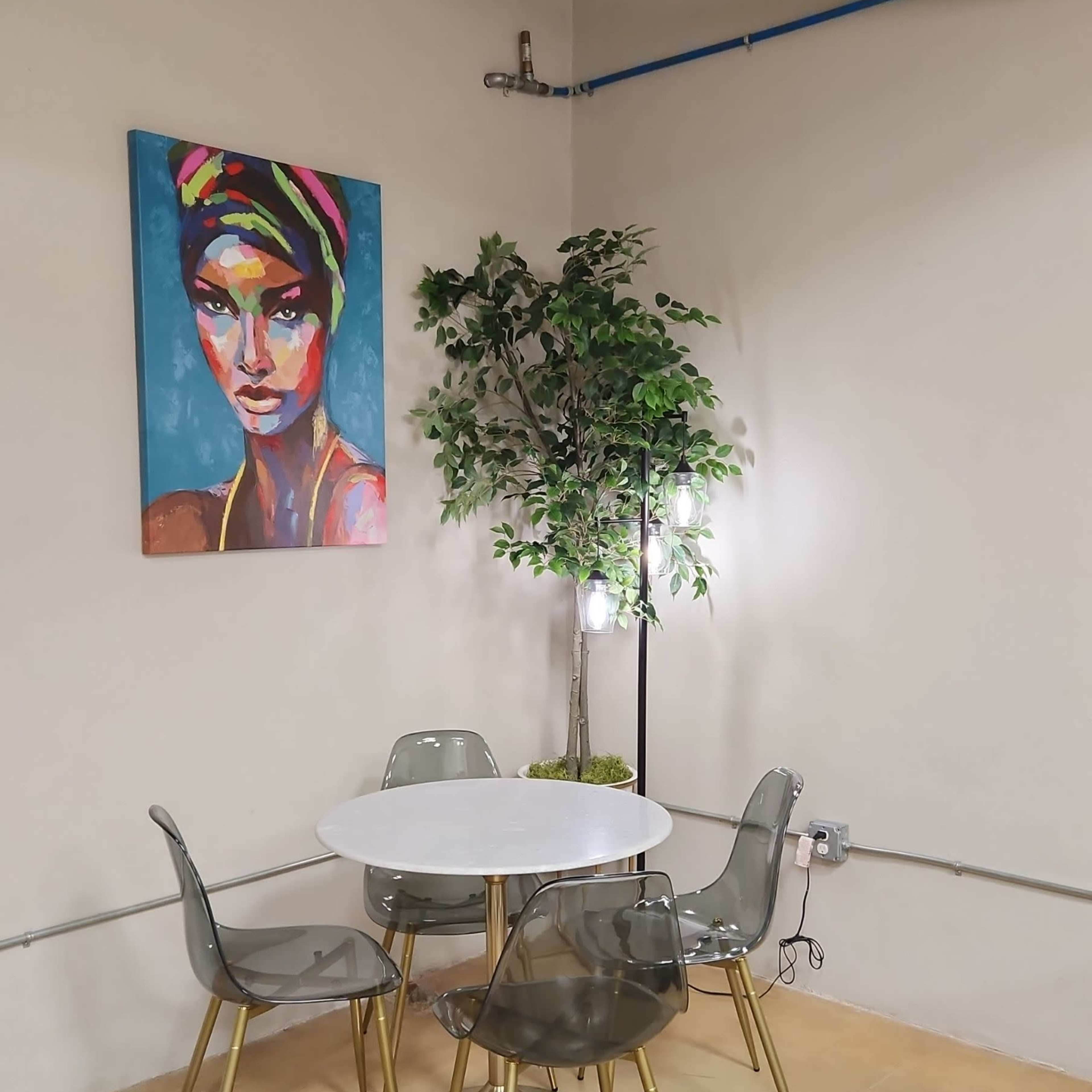 A small dining area features a round white table surrounded by transparent chairs, with a colorful portrait on the wall and a potted plant nearby.