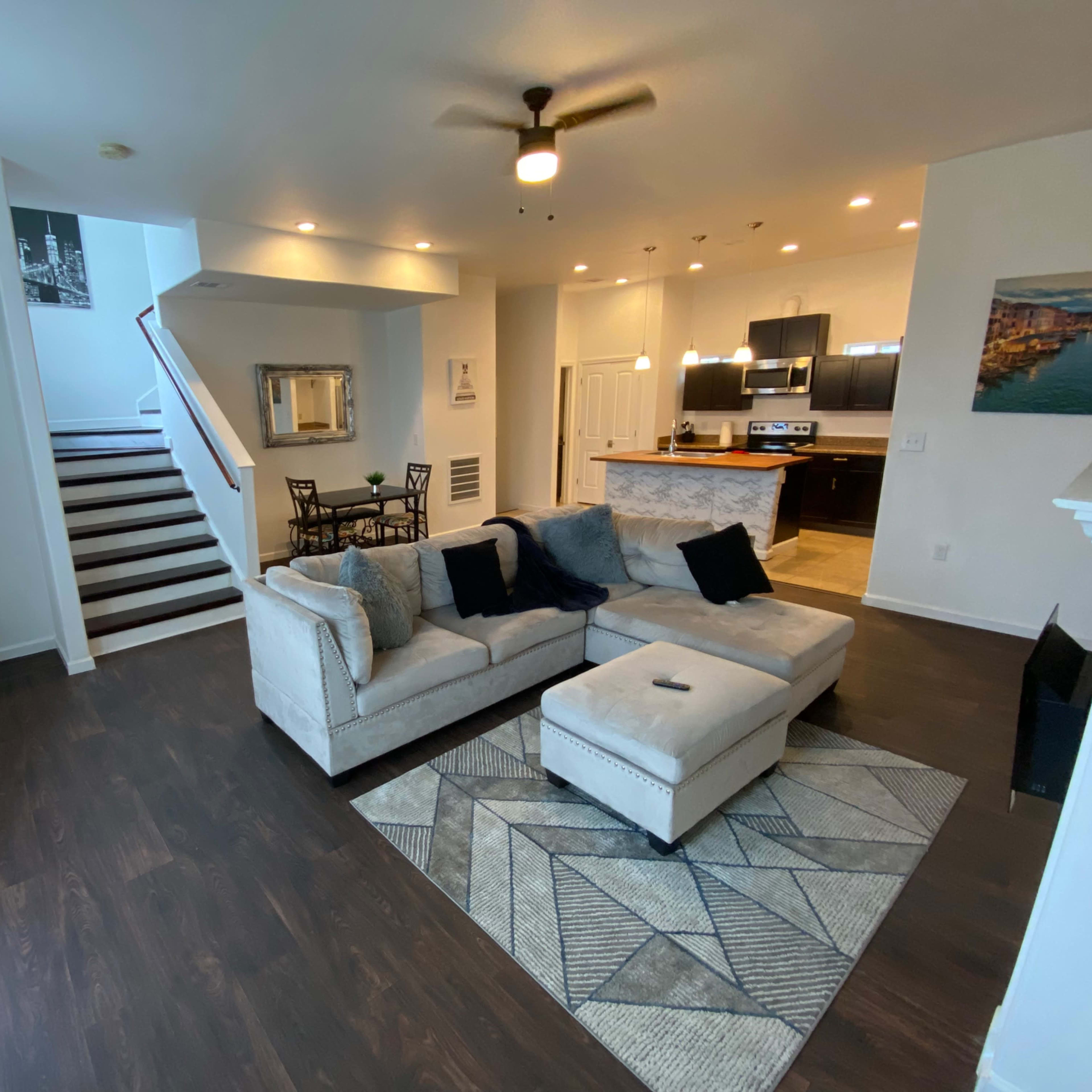 A modern living room features a light gray sectional sofa, a patterned area rug, and a staircase leading to an upper level.