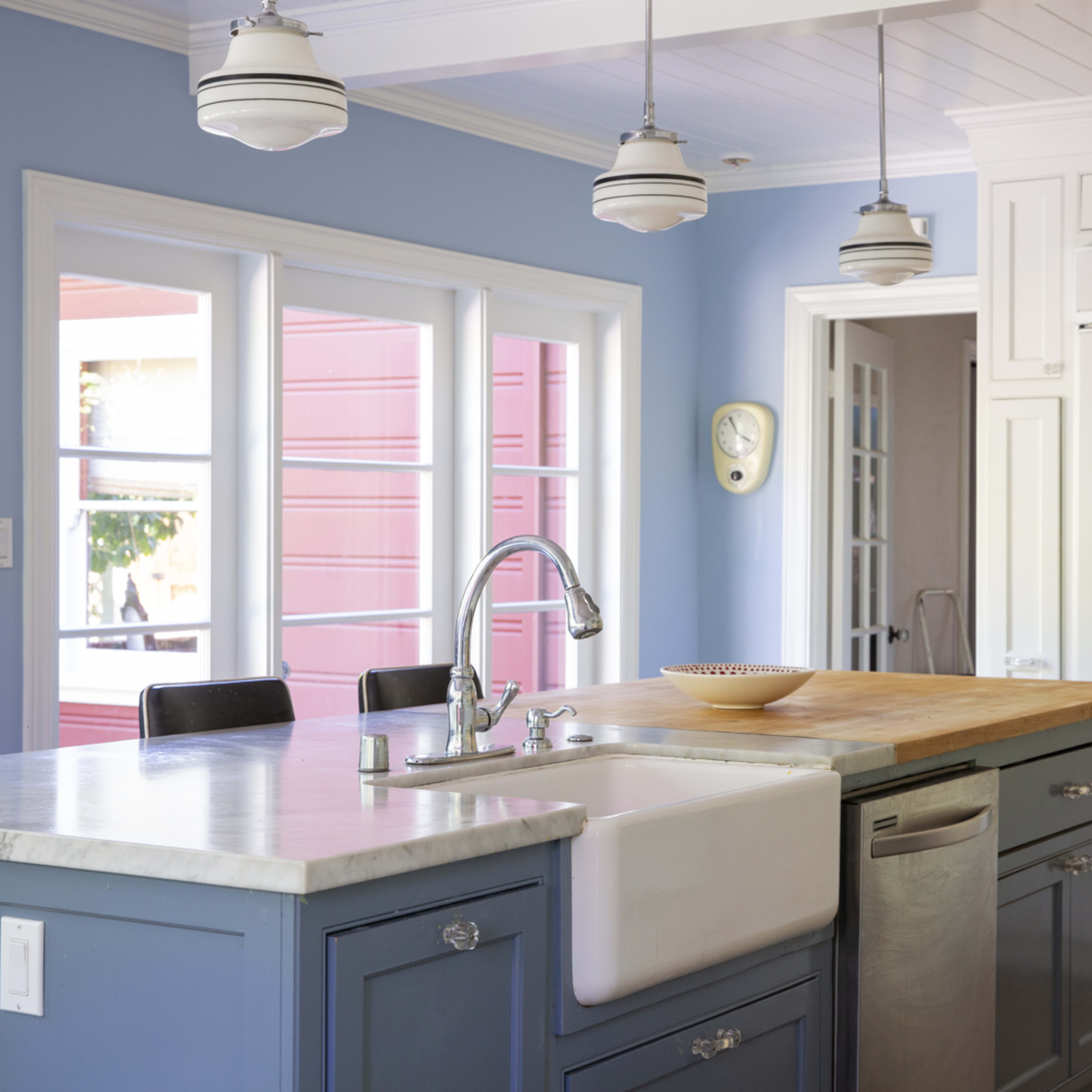 A kitchen features blue walls, a central island with a marble countertop and farmhouse sink, and large windows allowing natural light.