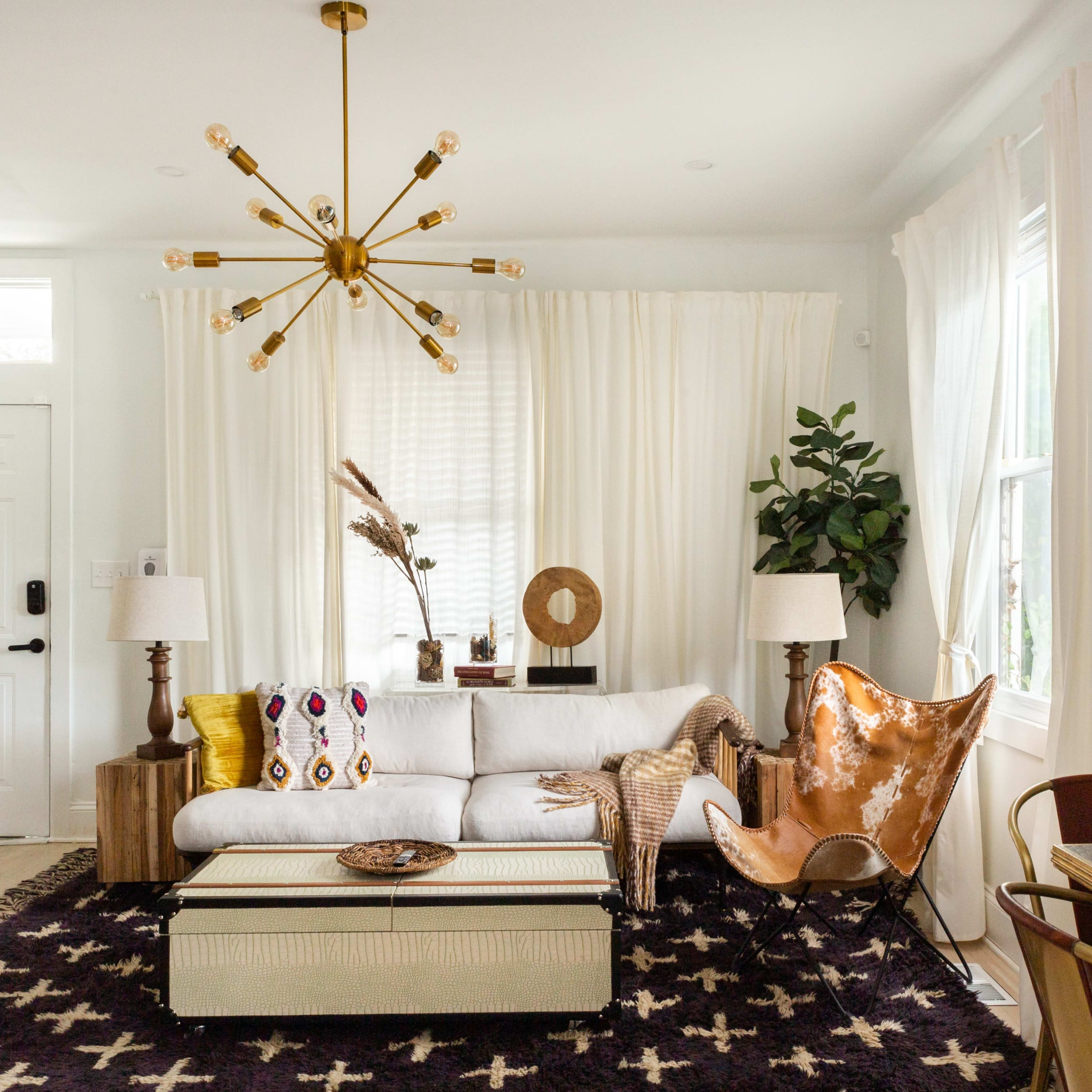 A modern living room features a white sofa, a decorative coffee table, a large black area rug, two lamps, and a leather chair, with potted plants and wall decor enhancing the space.