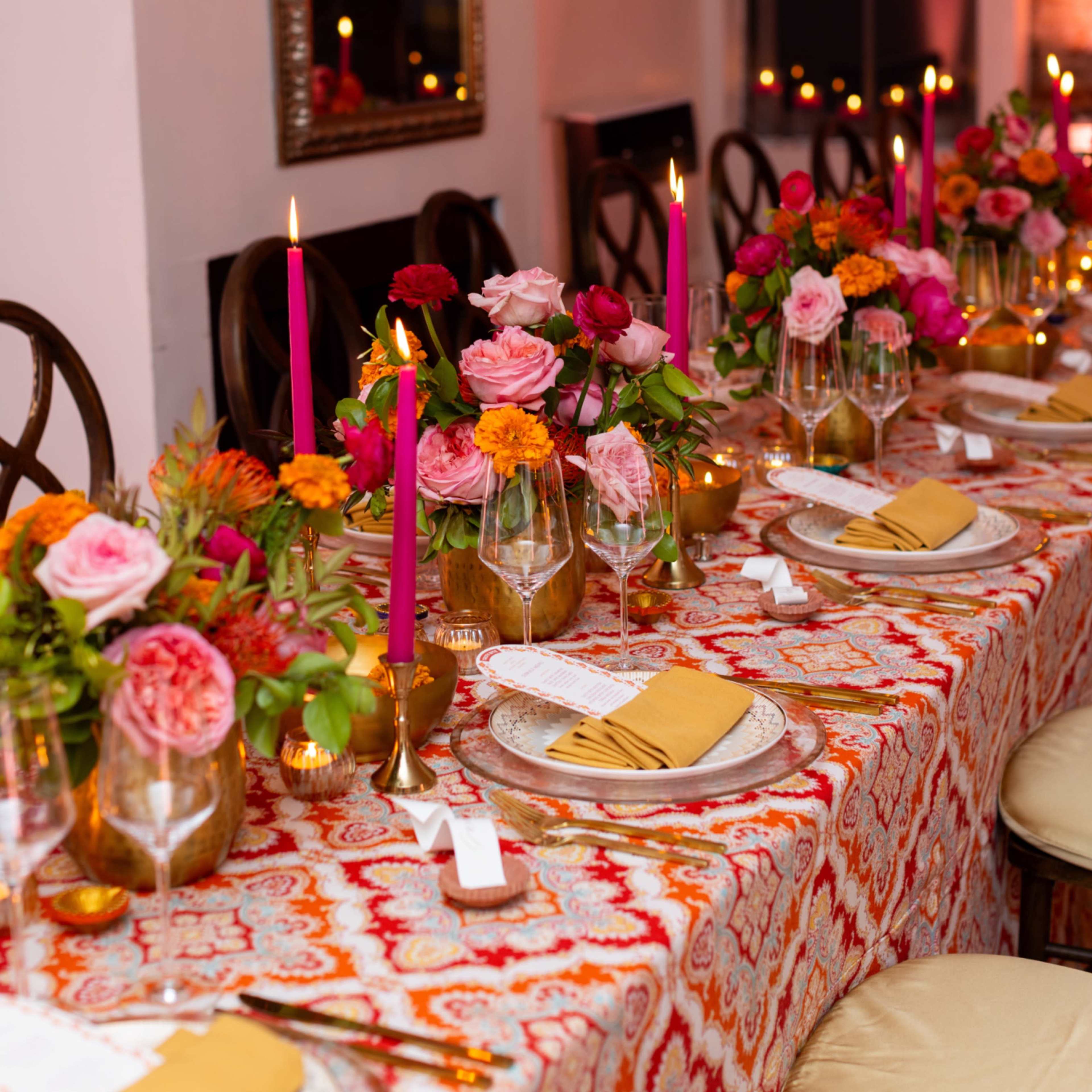 A long dining table is set with floral centerpieces, golden tableware, and pink candles, surrounded by chairs in a brightly lit room.