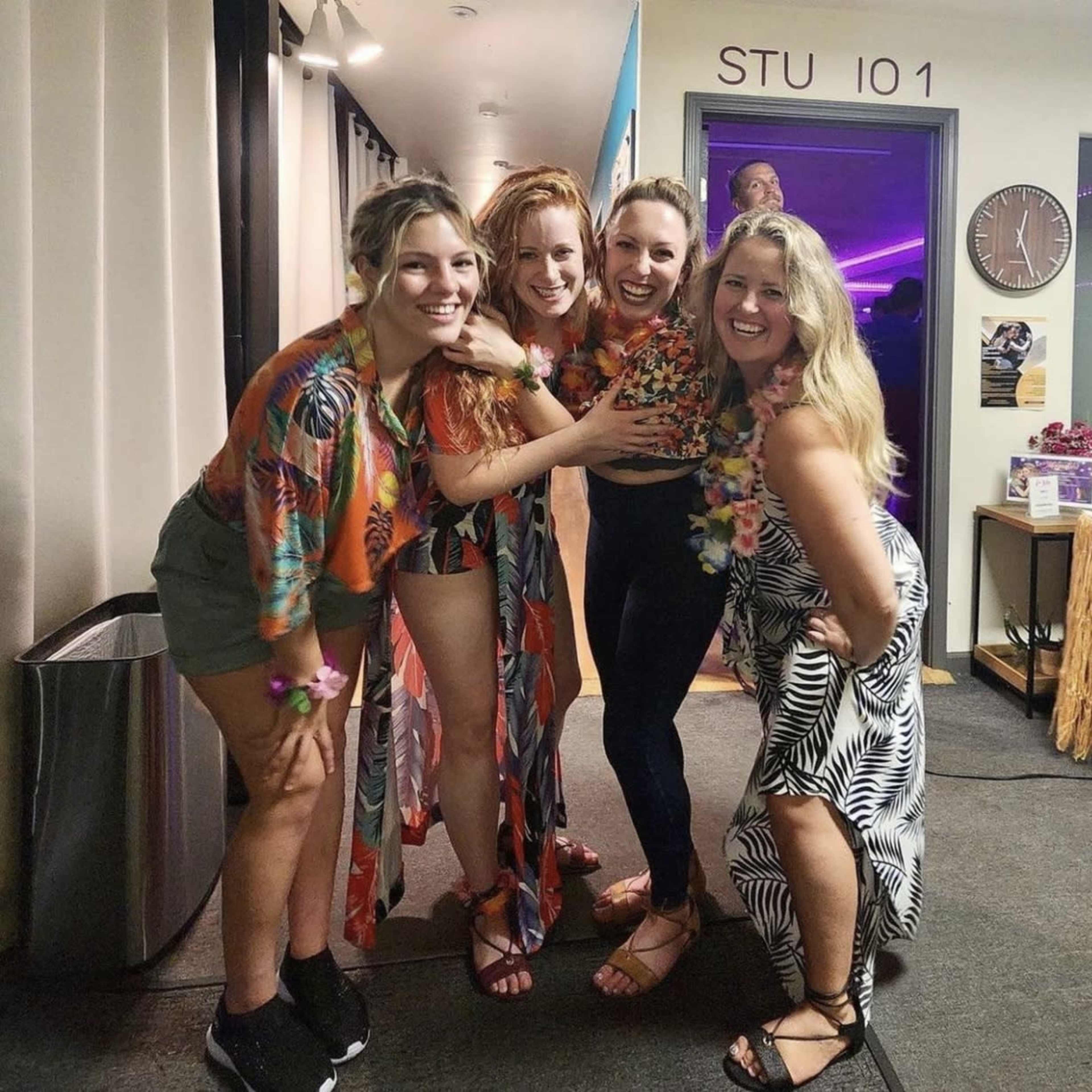 Four women stand together smiling, dressed in colorful clothing with floral patterns and leis, in a brightly lit indoor space.