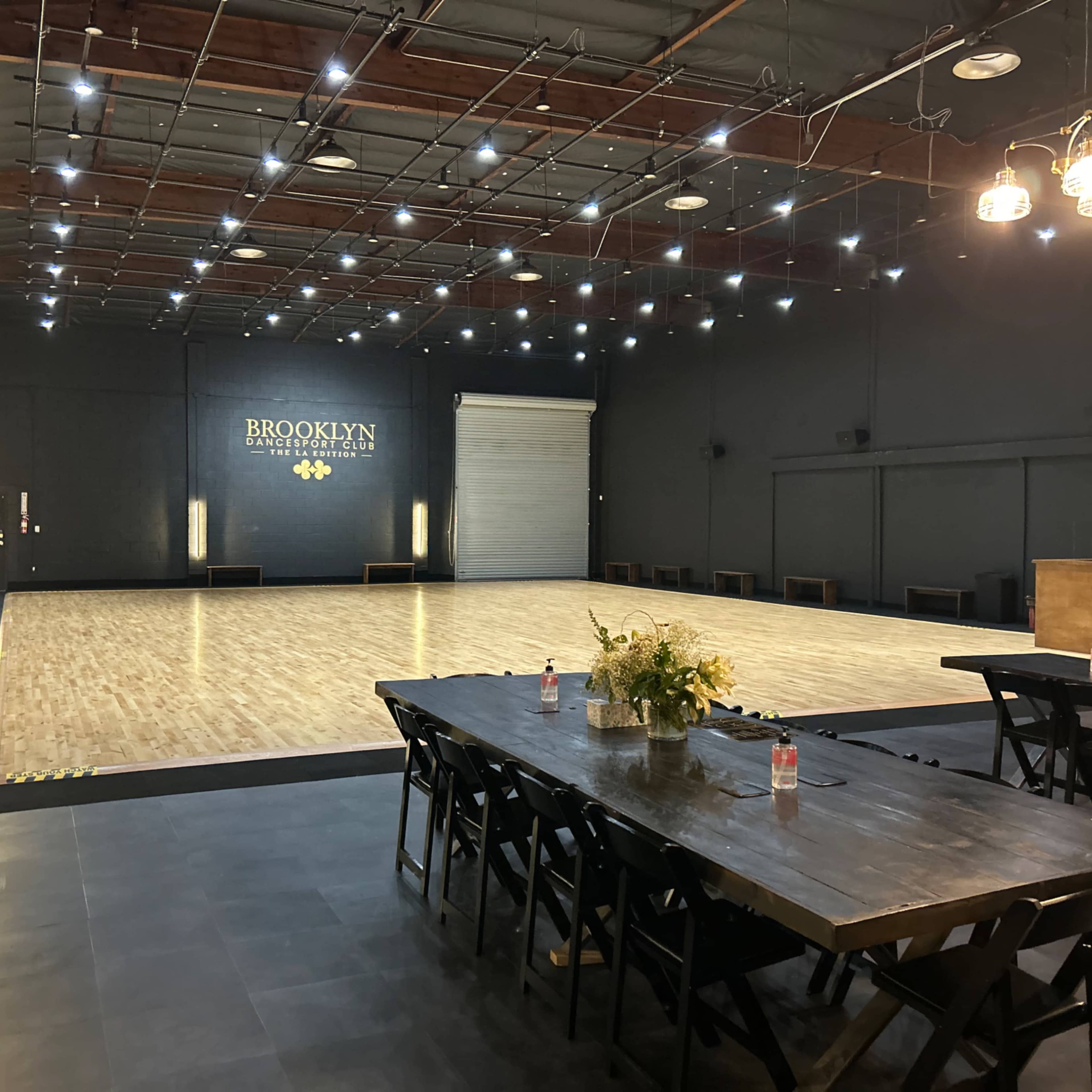 A spacious dance studio with a polished wooden floor, dim lighting, and a long table set for dining or gatherings along one side.