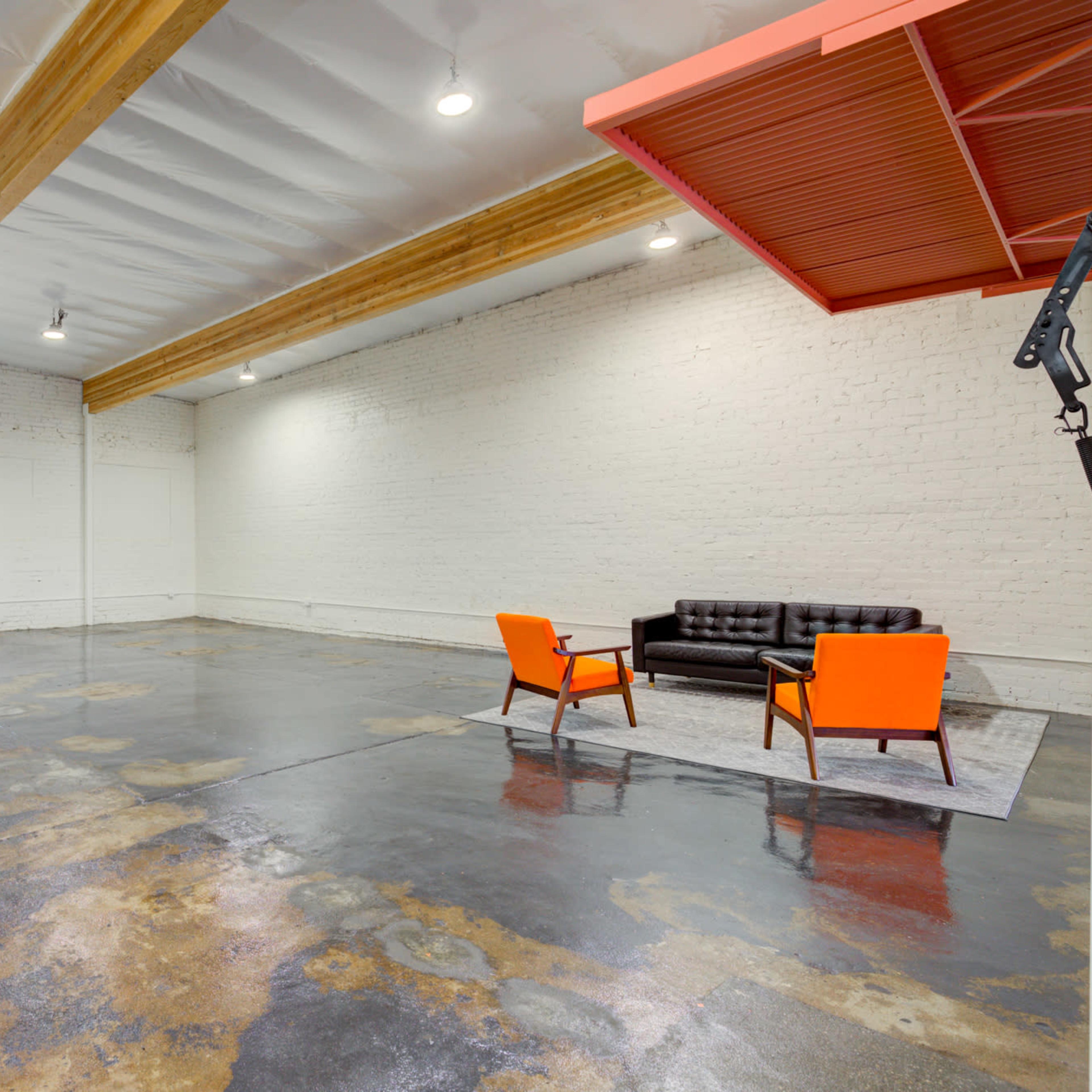 An empty room with a polished concrete floor, featuring a black leather couch and two orange chairs under exposed wooden beams and bright overhead lights.