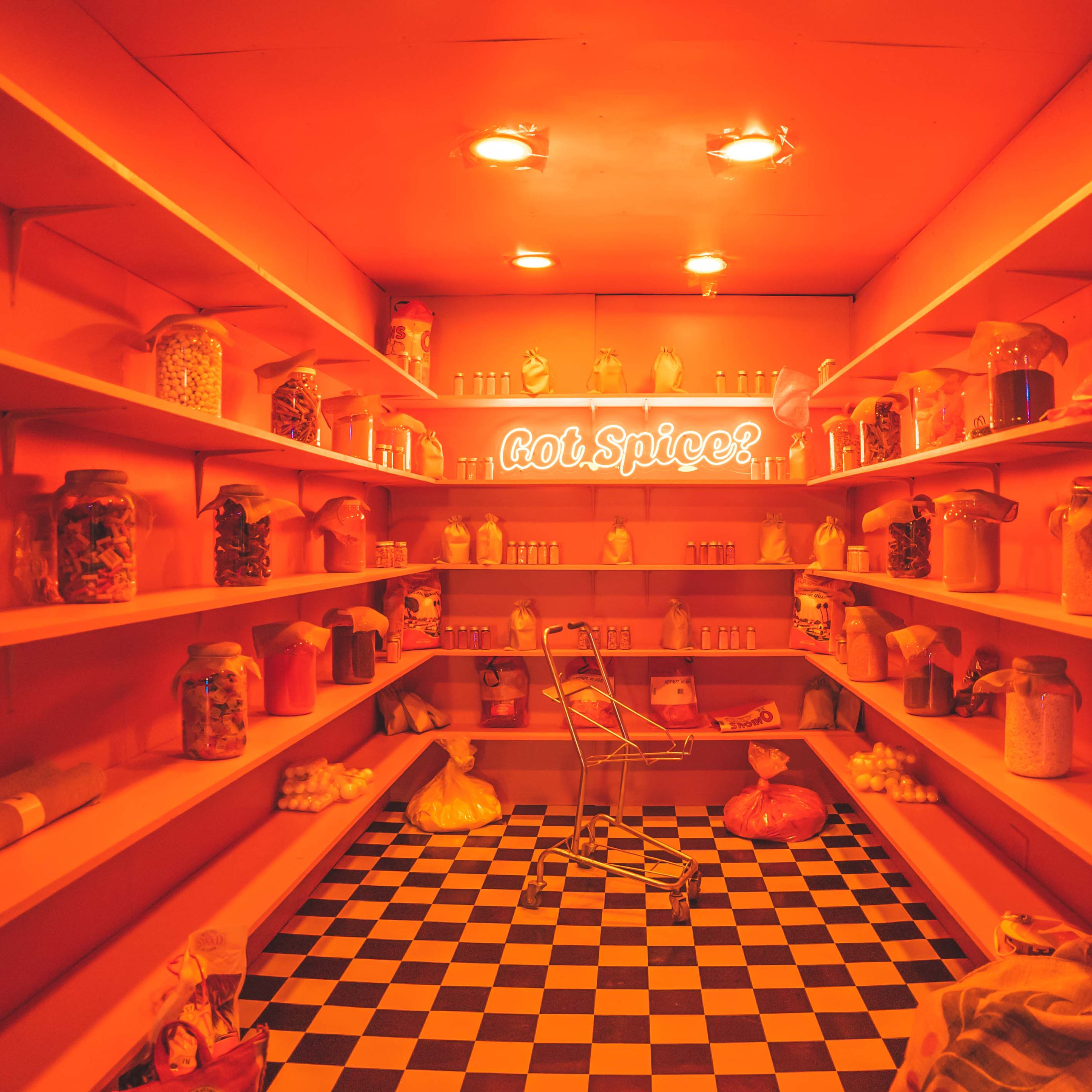 A brightly lit, orange-toned pantry features shelves lined with jars and containers of various ingredients, with a neon sign that reads "hot spice!" at the center.