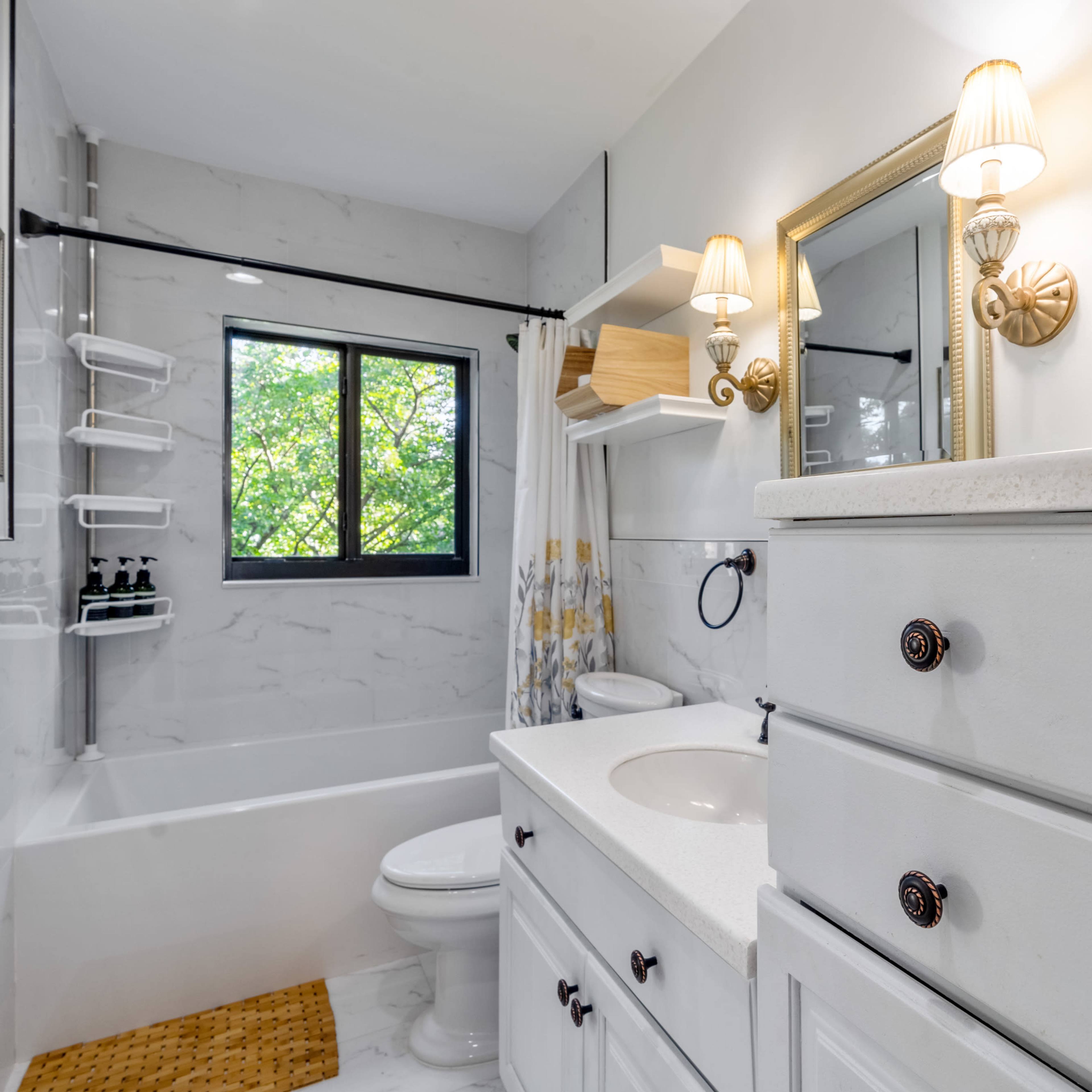 The bathroom features a white countertop, a bathtub with a shower, and natural light coming through a window, complemented by neatly arranged storage.