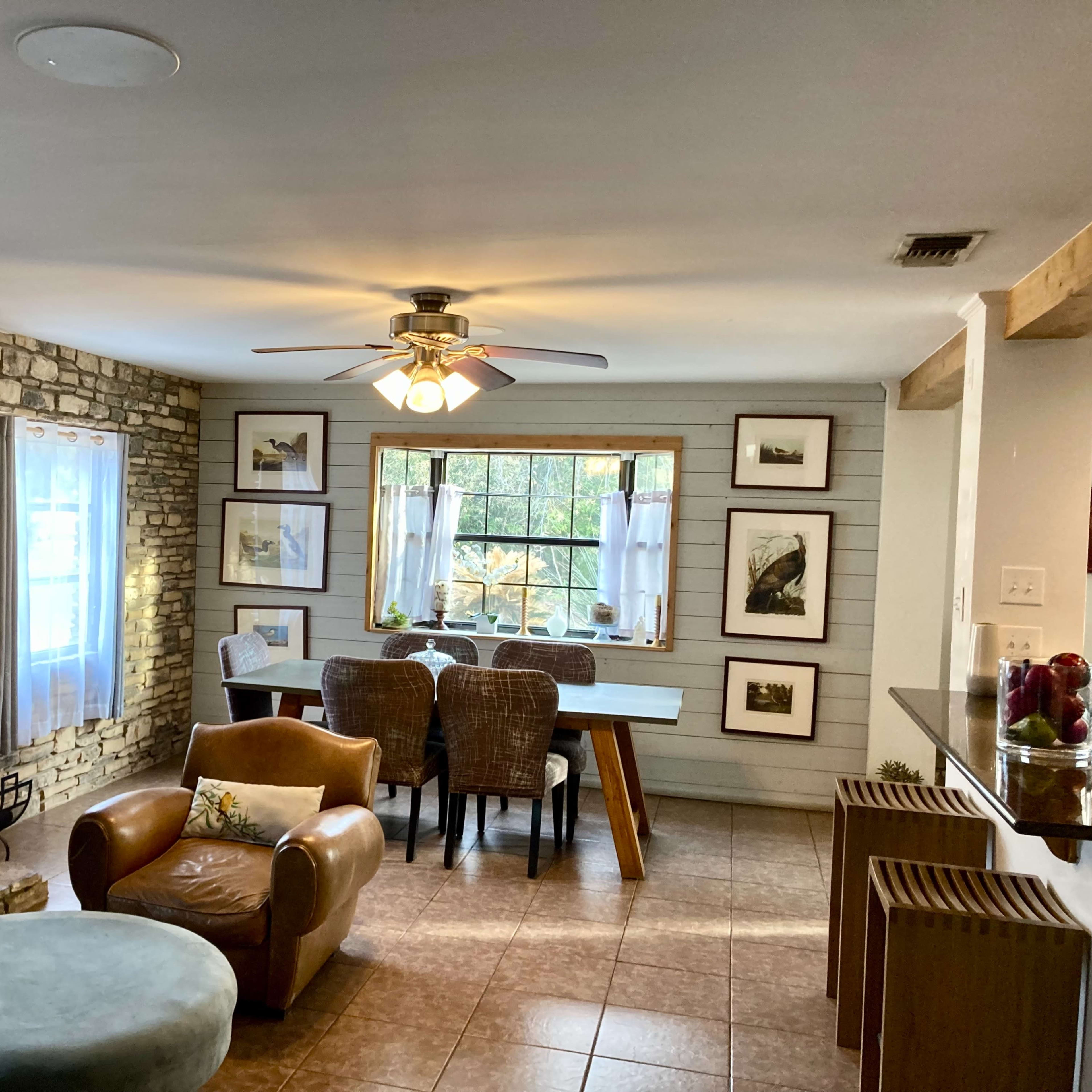 A cozy living space featuring a stone fireplace, a dining table with wicker chairs, and large windows with white curtains.