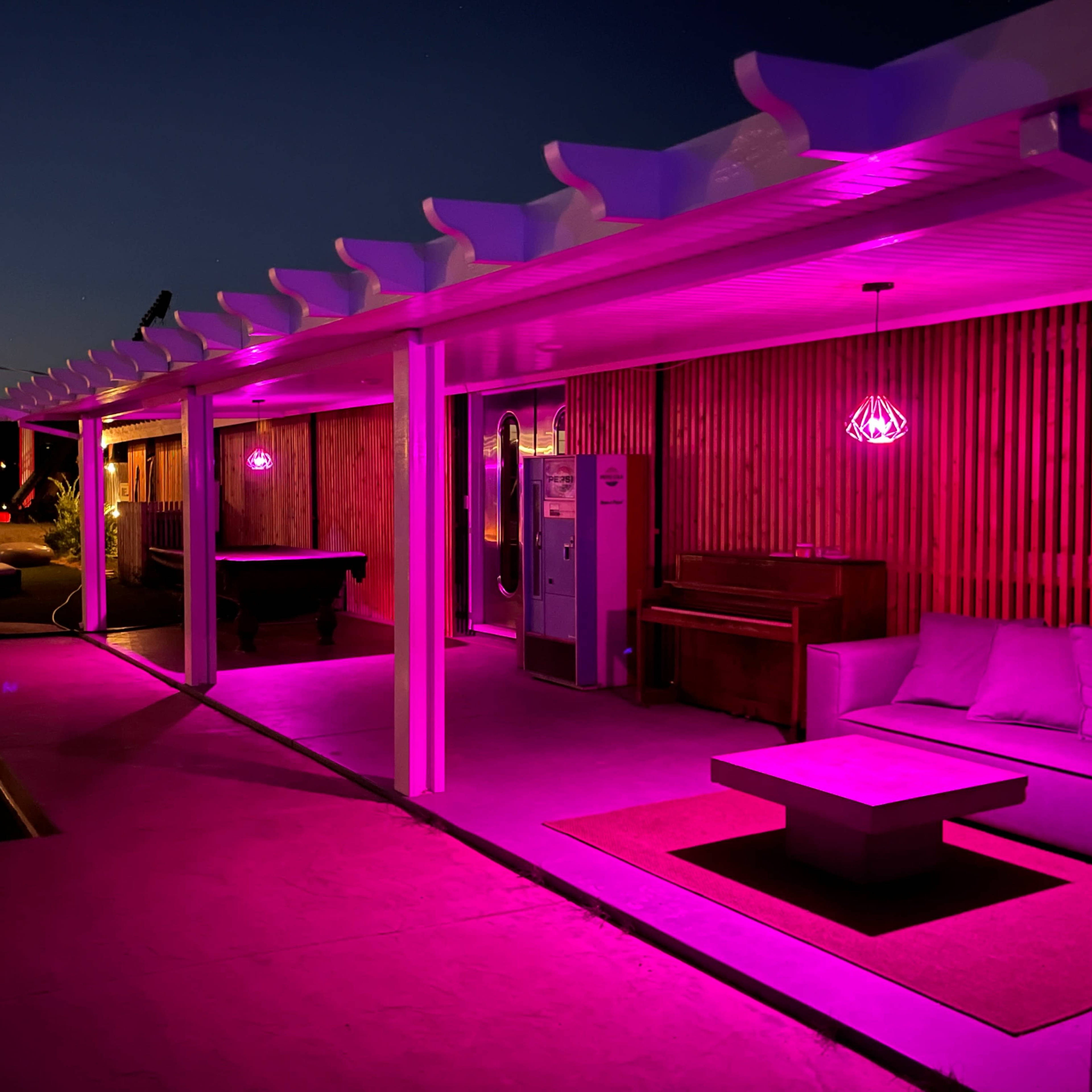 The scene features a modern outdoor lounge area illuminated by pink lights, with a pool, seating, and a piano under a covered structure.
