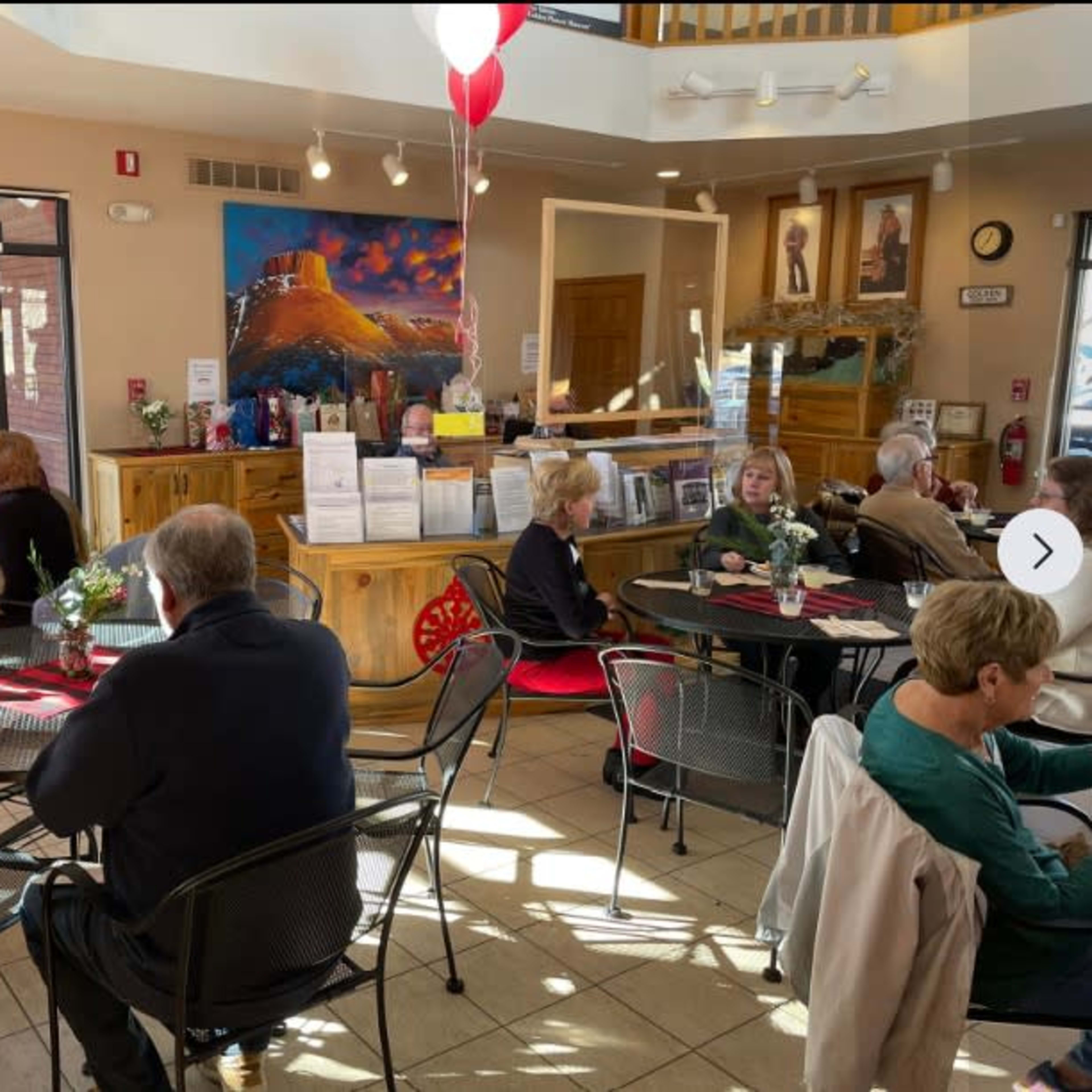 A crowded café with patrons sitting at round tables, engaging in conversation and enjoying their meals, while a large painting and framed artworks adorn the walls.