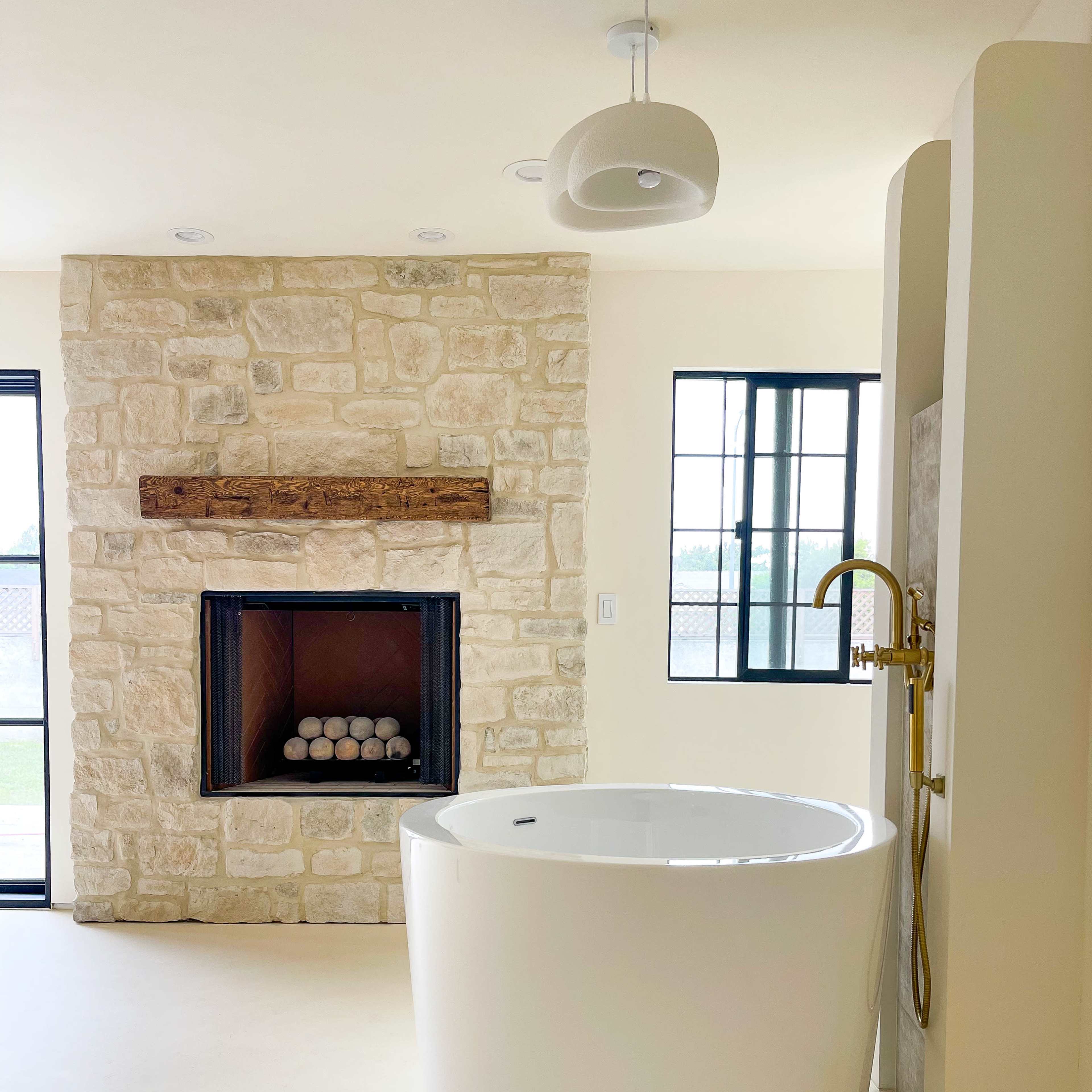 A modern bathroom featuring a freestanding round bathtub, a stone fireplace with a wooden mantel, and large windows allowing natural light.