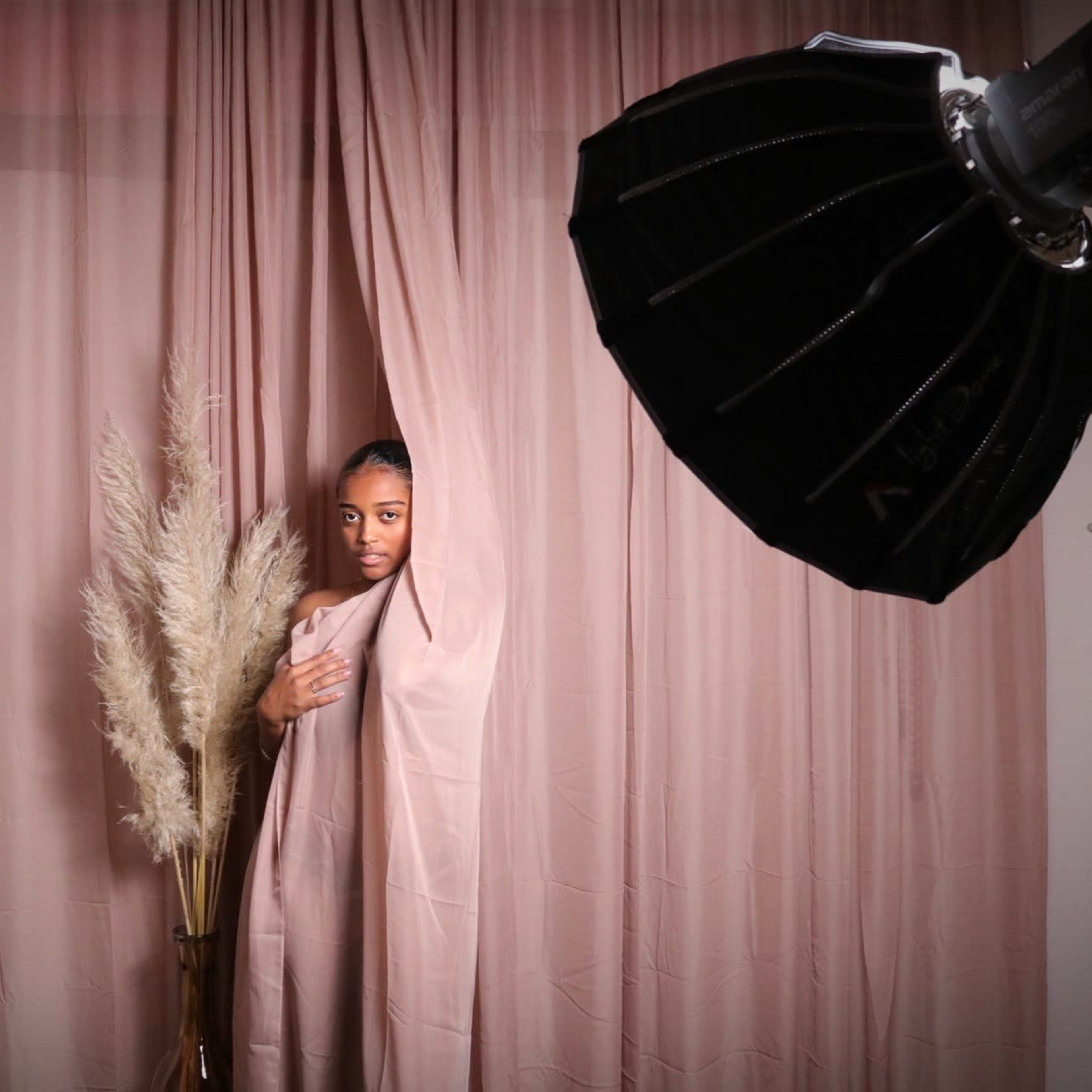 A person stands behind a flowing fabric in front of a soft pink backdrop, with a large photography light source and decorative pampas grass nearby.