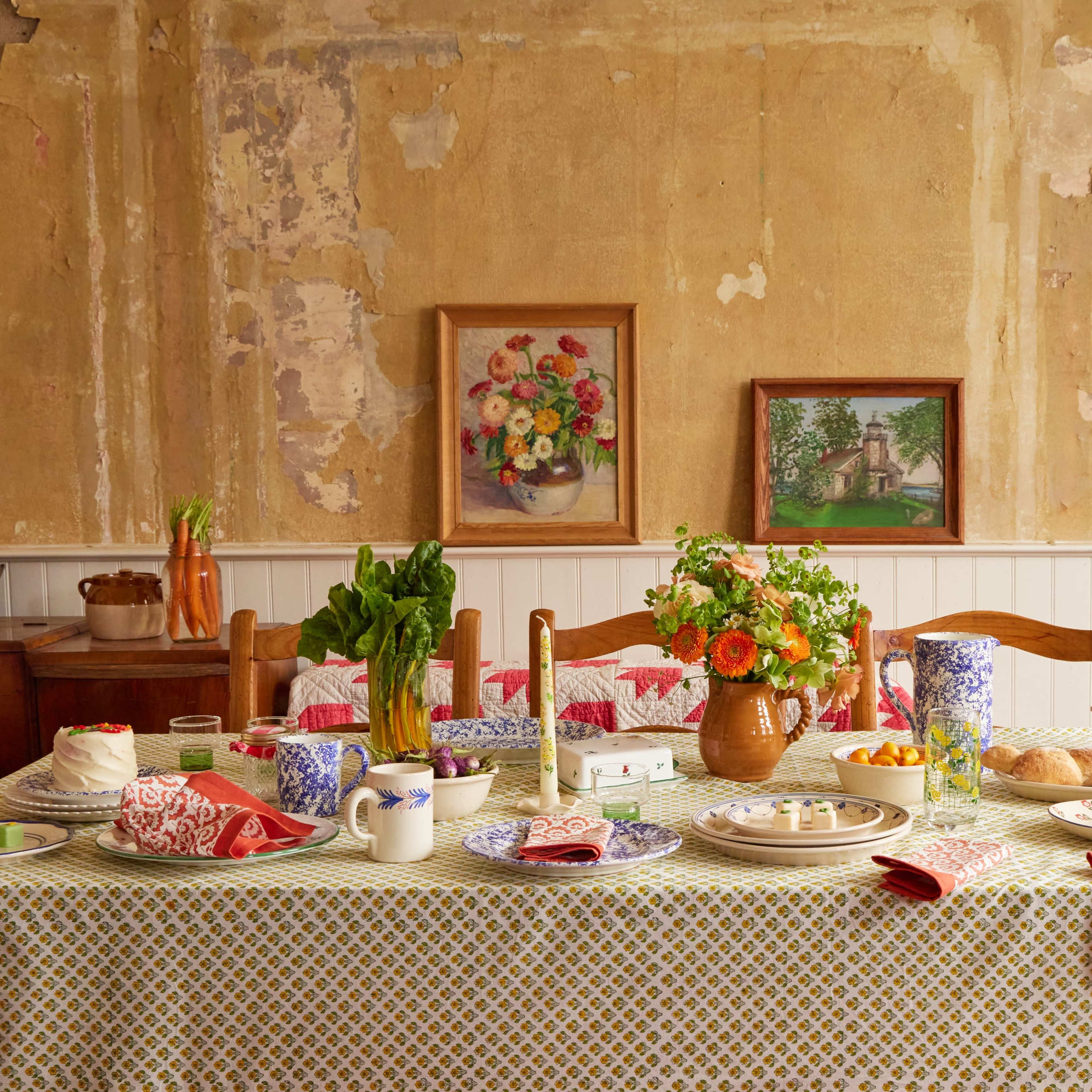 A dining table is set with various plates, a bouquet of flowers, and a spread of food in a rustic room with peeling walls and framed artworks.