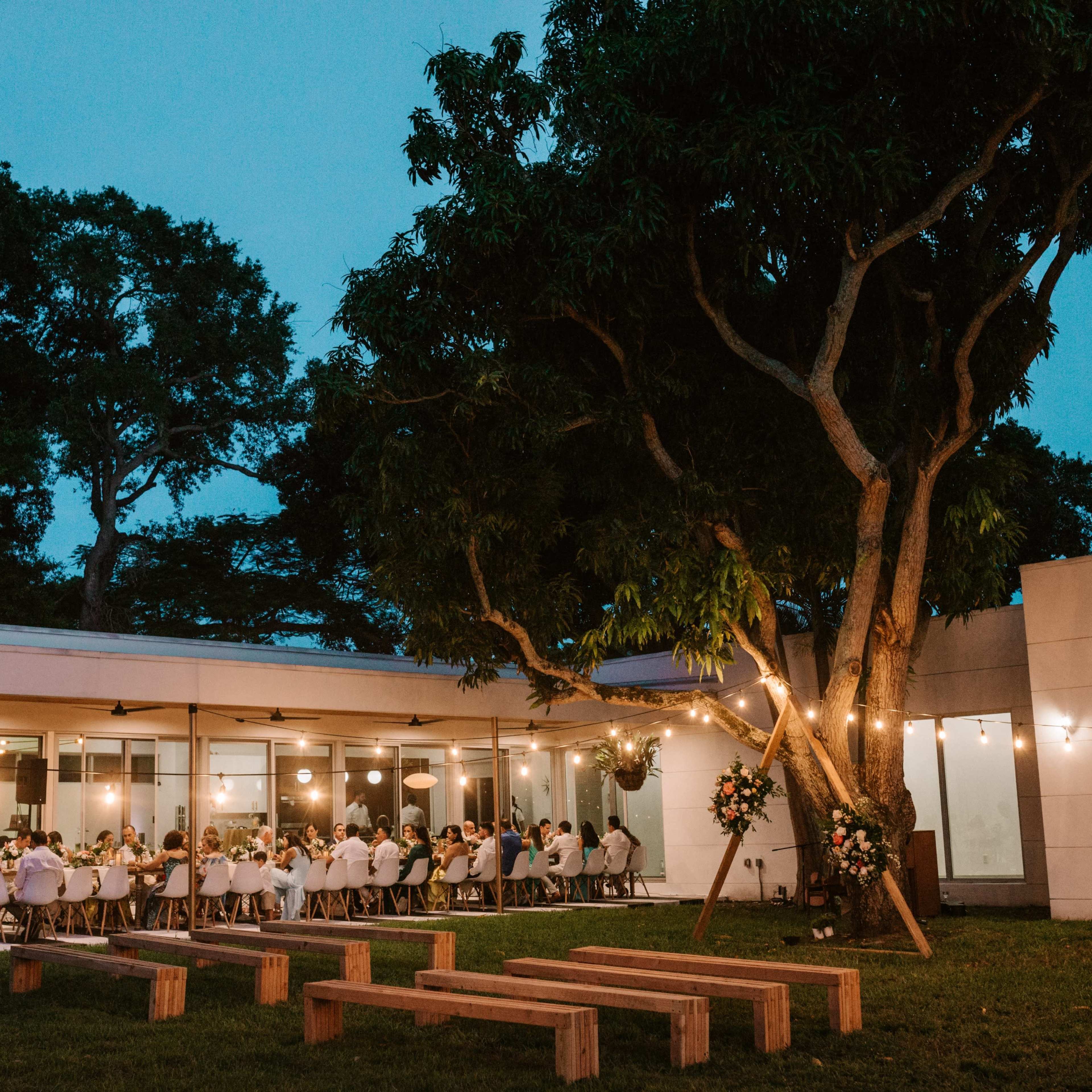 A large group of people dining outdoors under string lights next to a modern building surrounded by trees.